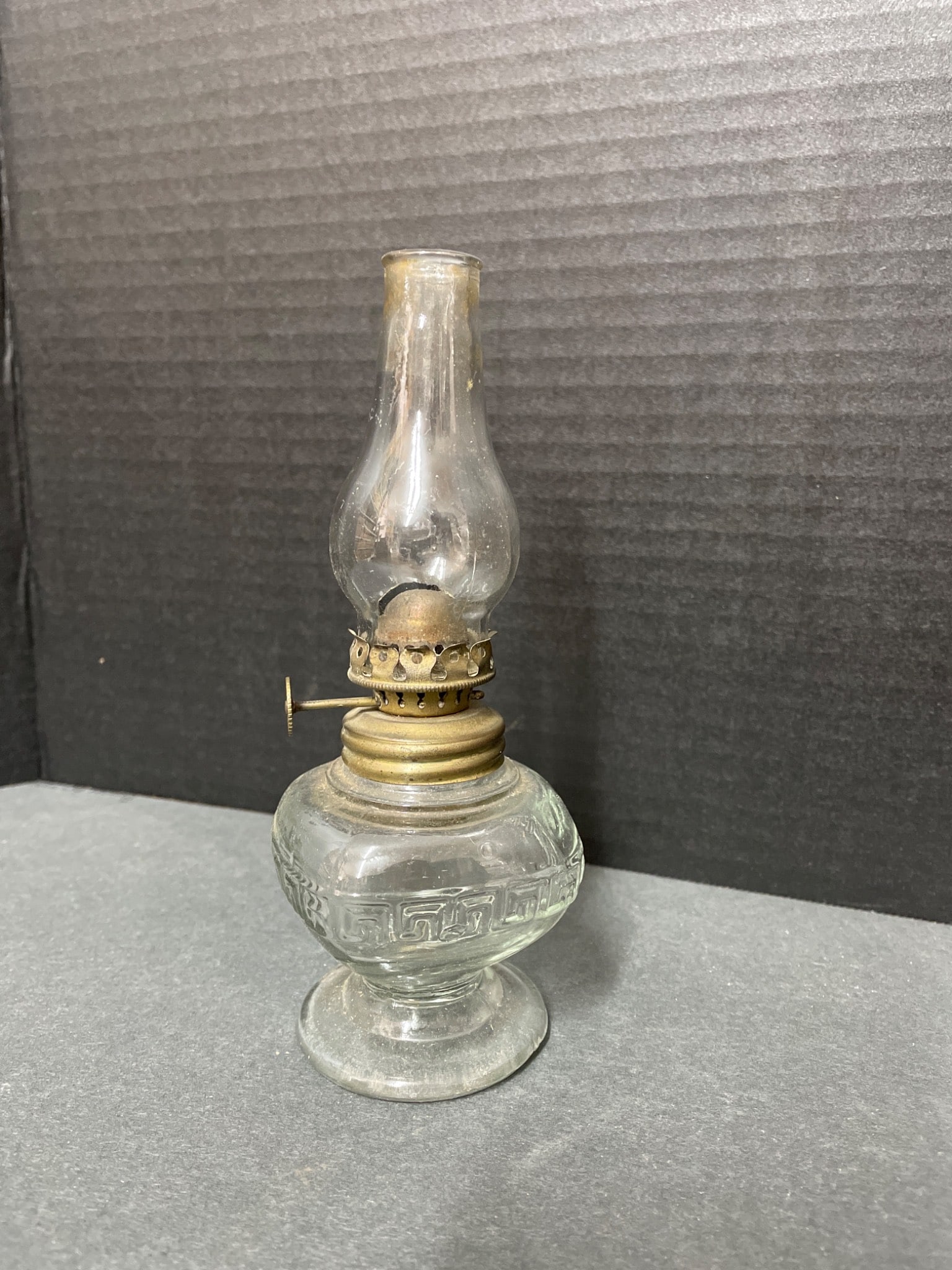 Glass Oil Lamp with Brass Burner and Glass Chimney: This is a glass oil lamp with a brass burner and detachable glass chimney. The lamp features a clear glass base with patterned detailing and a functional wick adjuster on the burner. All components,