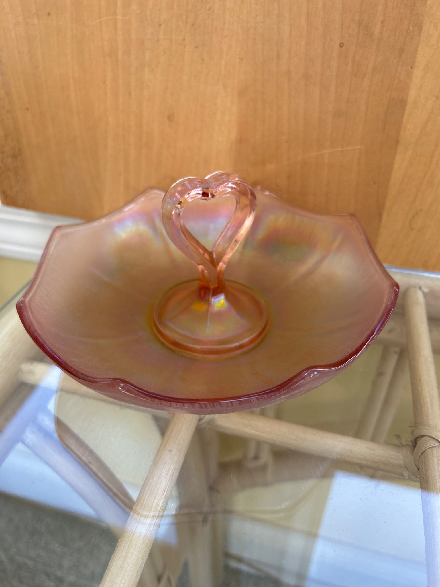 Footed Pink Iridescent Glass Candy Dish With Heart-Shaped Handle: This is a pink iridescent glass candy dish featuring a heart-shaped handle and a scalloped edge design. The dish displays a luster finish with multicolored reflections. It is likely made of pressed gl