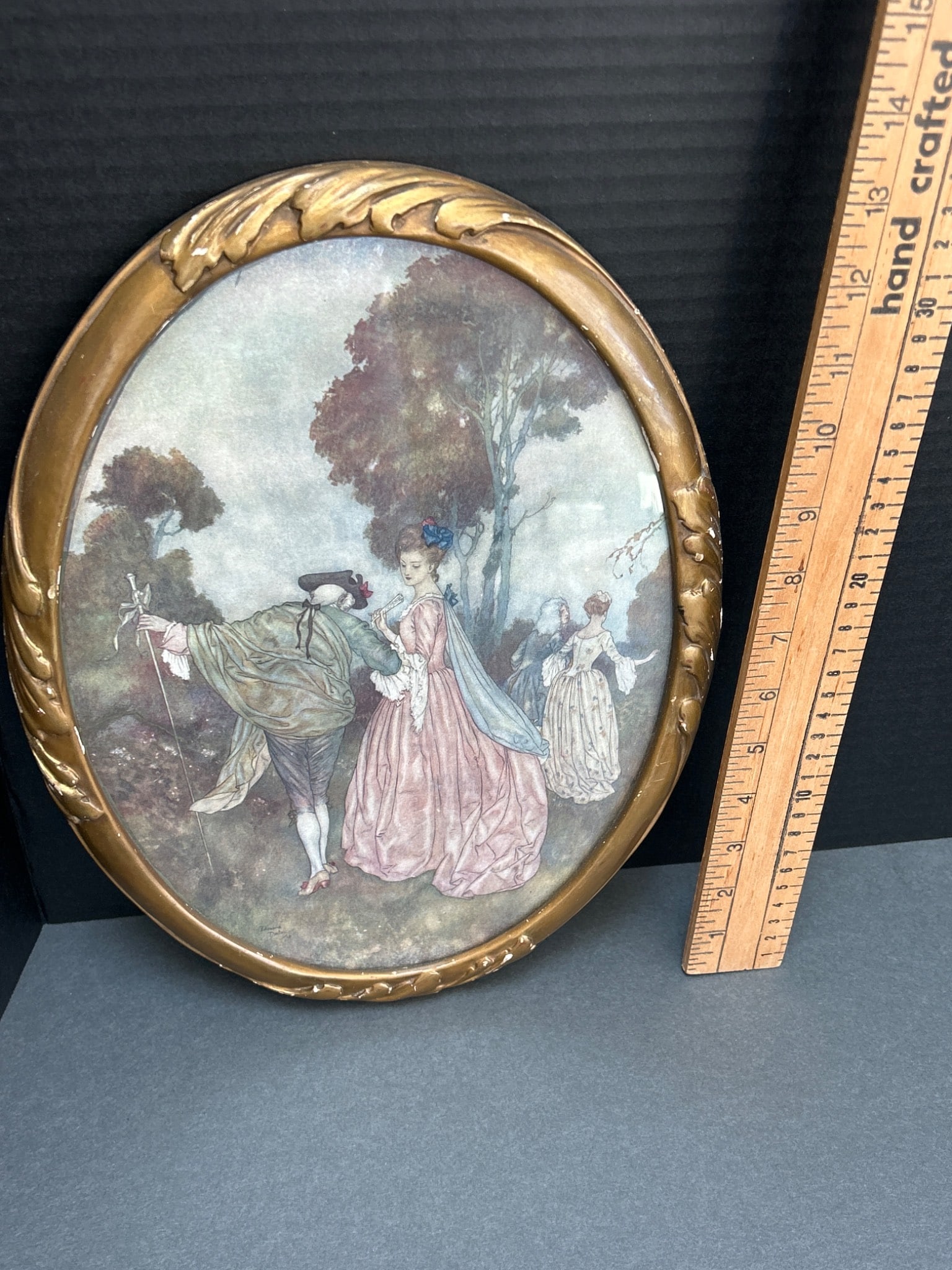 Oval Wall Art with Pastoral Scene in Gilded Frame: An oval-shaped decorative wall art piece featuring a pastoral scene with figures in period attire. Encased in a gilded frame with a leaf motif. Likely a print on paper or board. The mount and backer b