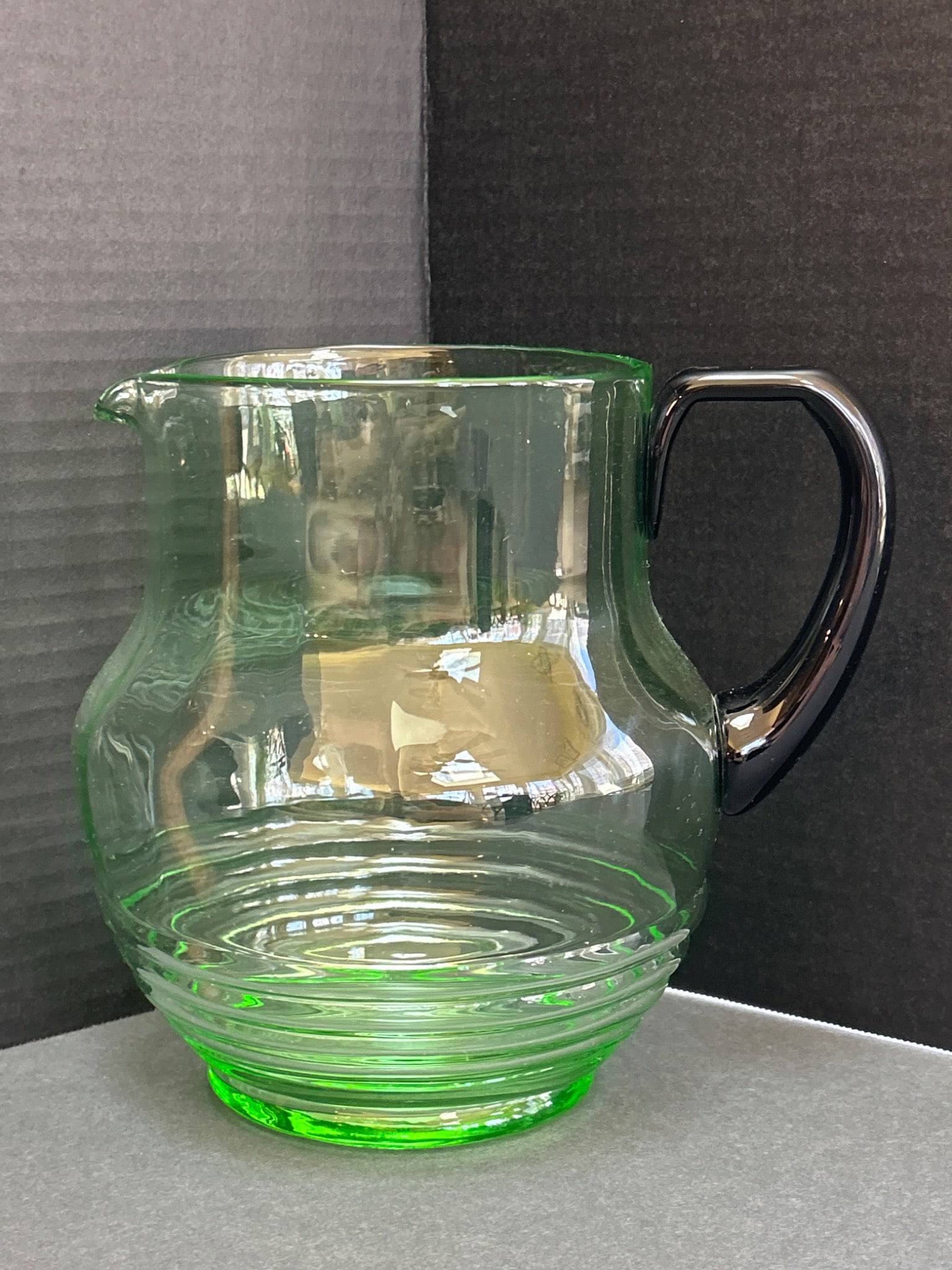 Glass Pitcher With Black Handle and Green Body: Glass pitcher featuring a green round-ribbed body and a contrasting black handle. The pitcher includes a spout for pouring and measures approximately 6 inches in height. Its lightweight, transparent