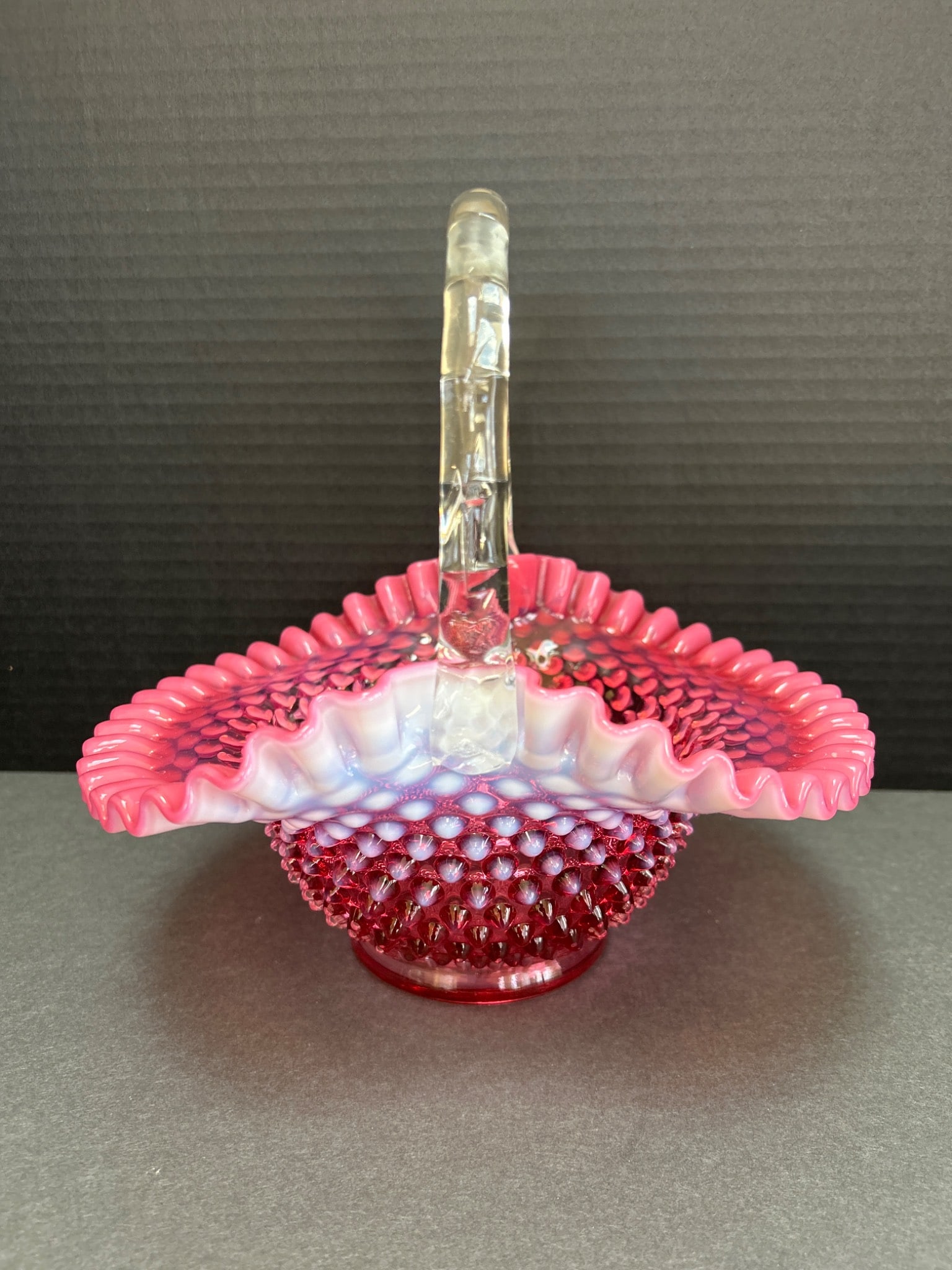 Ruby Red Hobnail Glass Basket With Applied Handle: This is a ruby red hobnail glass basket featuring a clear applied glass handle. The basket has a ruffled edge design, with the primary color transitioning between pink and white near the edges. The ba