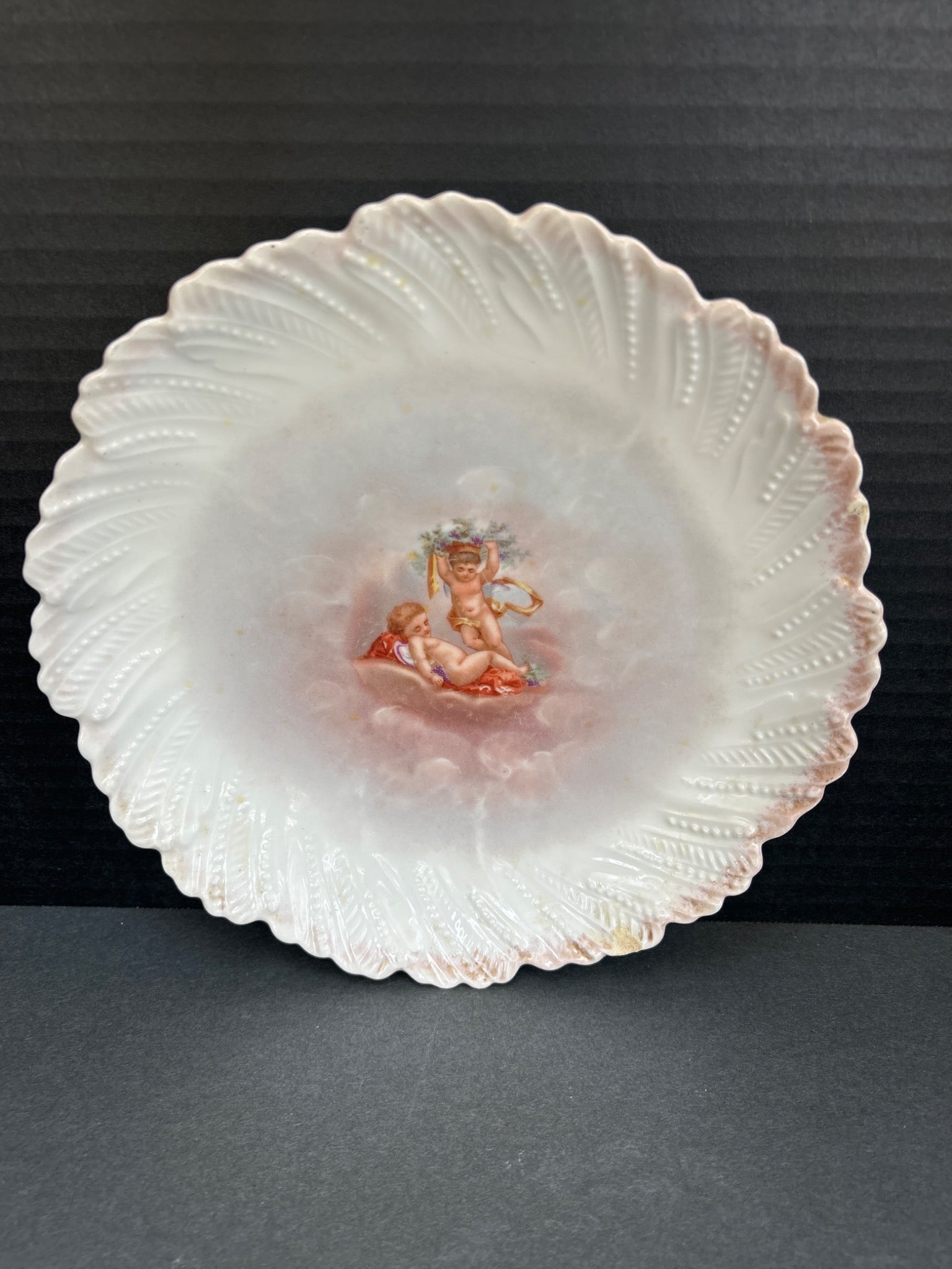 Limoges France Decorative Porcelain Plate With Cherub Design: This is a decorative porcelain plate marked 'Limoges France' on the back with an additional hallmark. It features a lithographed cherub design at the center and scalloped edges with embossed details.