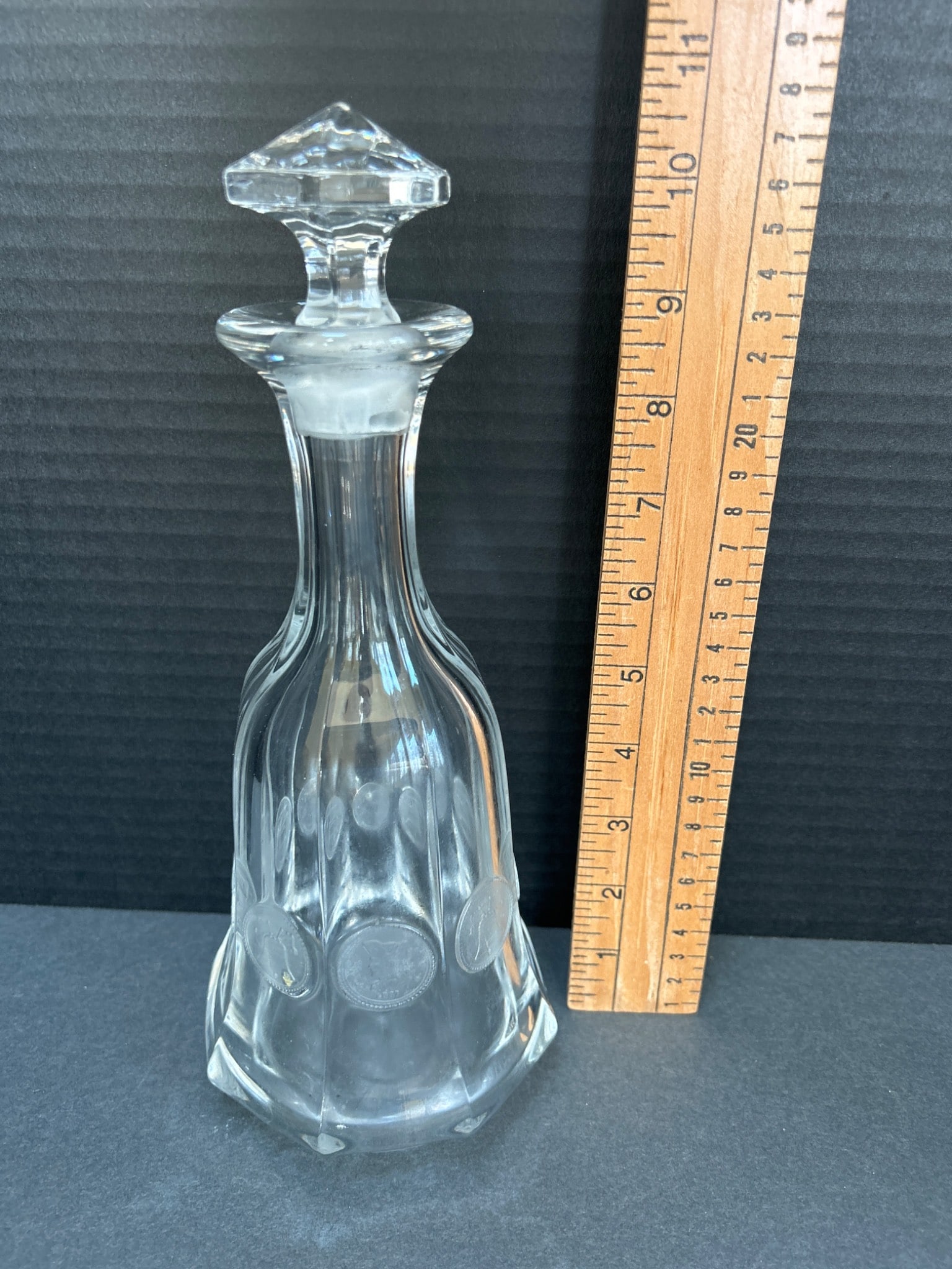 Clear Glass Decanter with Etched Coin Designs and Stopper: A clear glass decanter featuring etched coin motifs around the body. The decanter is approximately 11 inches tall with its stopper and features a hexagonal base. The stopper is cut, matching the