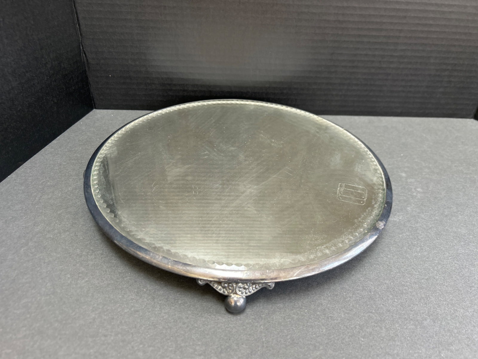 Round Mirror Tray With Metal Frame and Three-Legged Base: This is a round mirror tray featuring a beveled edge and mounted in a metal frame. It rests on a three-legged base with detailed ornamental feet. The diameter of the tray is approximately 12 inches. T