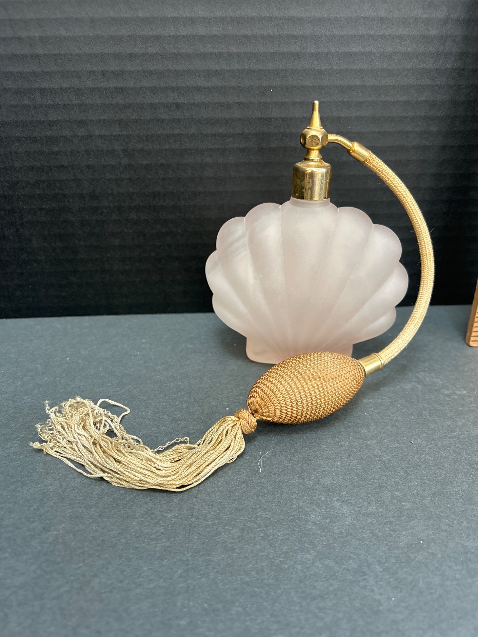 Glass Perfume Atomizer With Brass Components And Tassel Applicator: This item is a frosted glass perfume atomizer with a shell-shaped body. It features brass hardware, including a nozzle and neck, with an attached flexible hose and a fabric-covered pump applicator wit