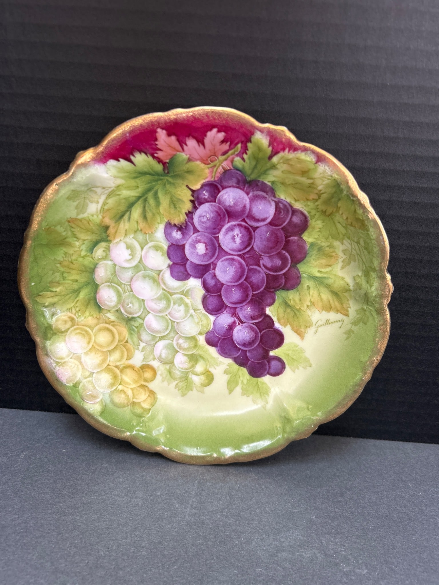 Bavarian Porcelain Plate with Grape Motif and Gold Trim: A decorative porcelain plate featuring hand-painted grape clusters in purple, white, and green with a scalloped, gold-trimmed edge. Marked on the back with a crown and shield insignia and 'Bavaria,'