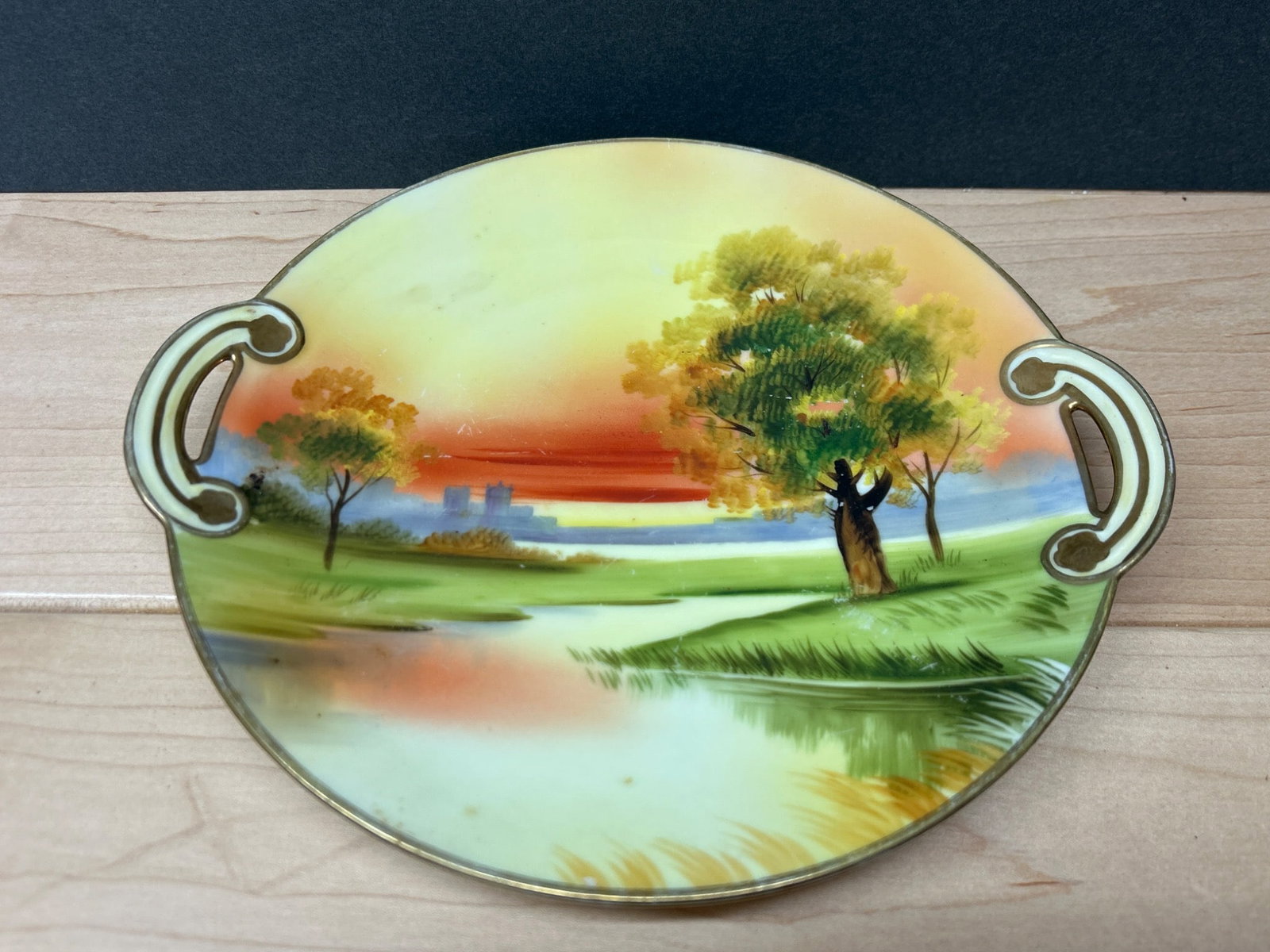 Hand-Painted Nippon Plate with Handles and Landscape Scene: Hand-painted porcelain plate featuring a landscape scene with trees, a river, and sunset tones. The plate includes two open-style handles and measures approximately 10 inches across, including the