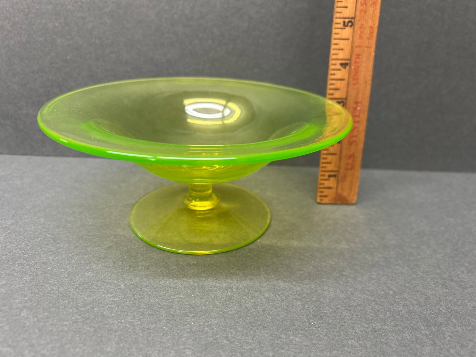 Uranium Glass Compote Dish with Pedestal Base: A uranium glass compote dish with a pedestal base. The dish measures approximately 4 inches in height, as shown against a ruler for reference. The greenish-yellow glow is characteristic of uranium