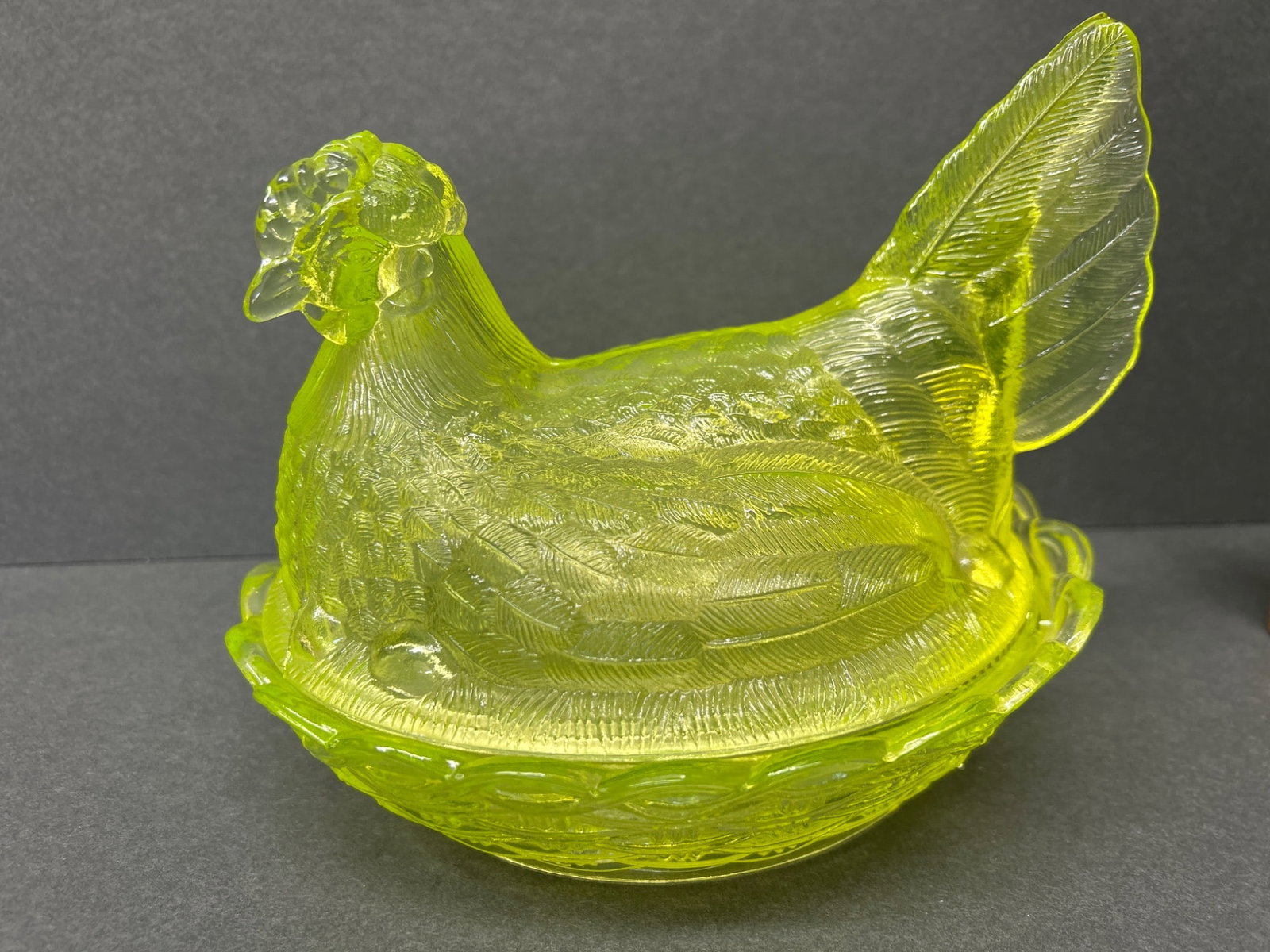 Pressed Uranium Glass Hen on Nest Covered Dish: This item is a pressed uranium glass dish in the shape of a hen on a nest. Typically used as a covered dish or collectible, it exhibits high detail with the hen acting as the lid. The dish emits a gre