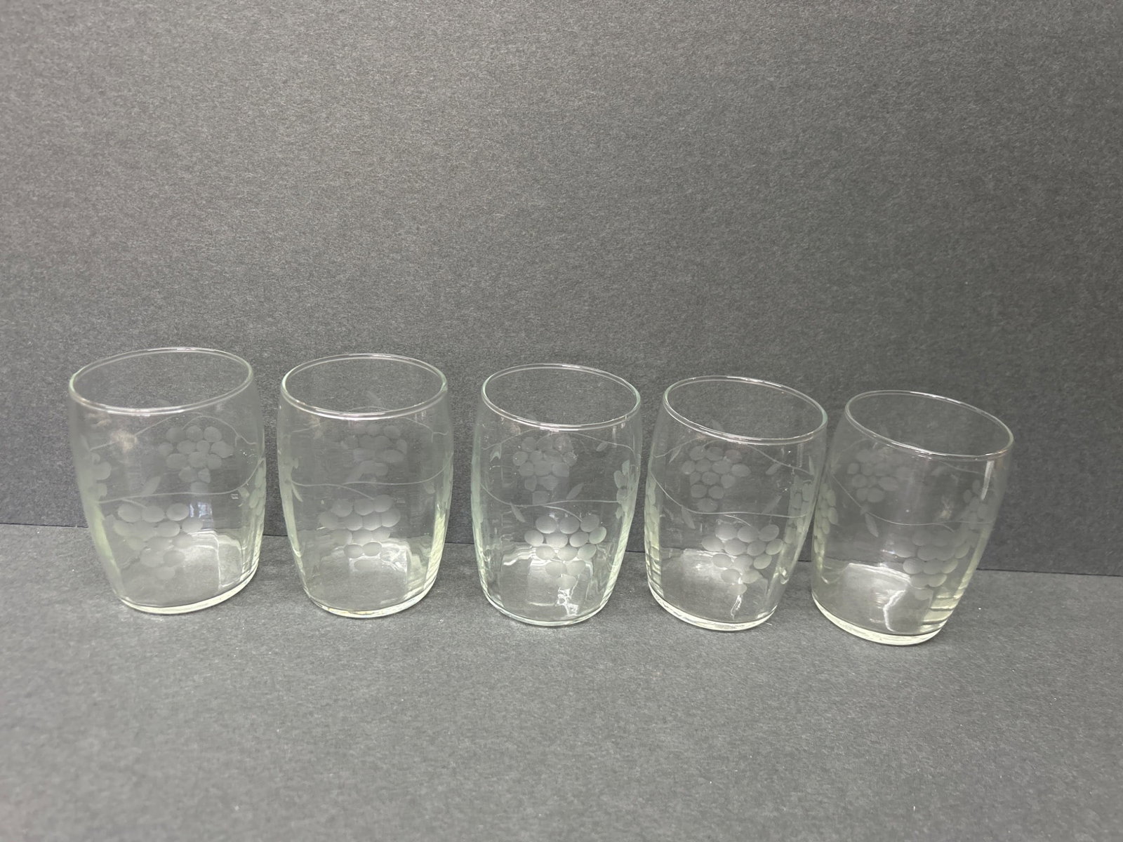5 Etched Glass Tumblers With Grape Design: 5 clear glass tumblers featuring an etched grape cluster pattern. Each glass measures approximately 4 inches in height with a 3-inch diameter. No visible branding or markings. Condition appears good w