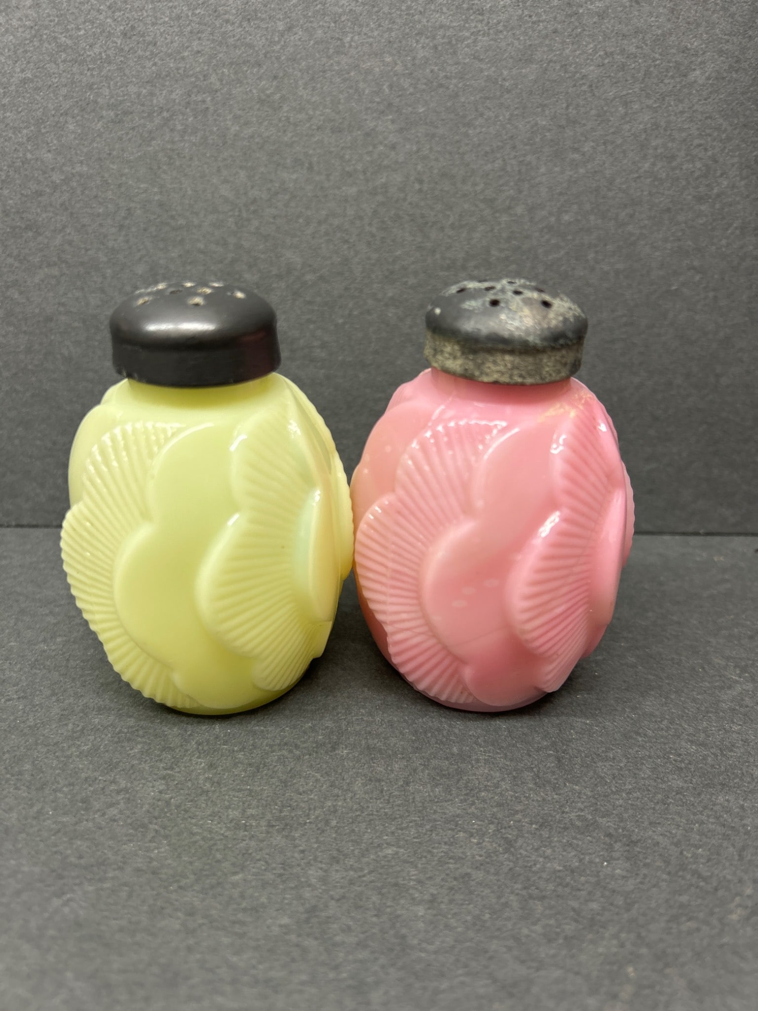 Pink and Yellow Pressed Glass Salt and Pepper Shakers With Metal Lids: Pressed glass salt and pepper shakers, one pink and one yellow, designed with a scalloped pattern. Both shakers are fitted with metal screw-on lids showing signs of wear and corrosion. The items