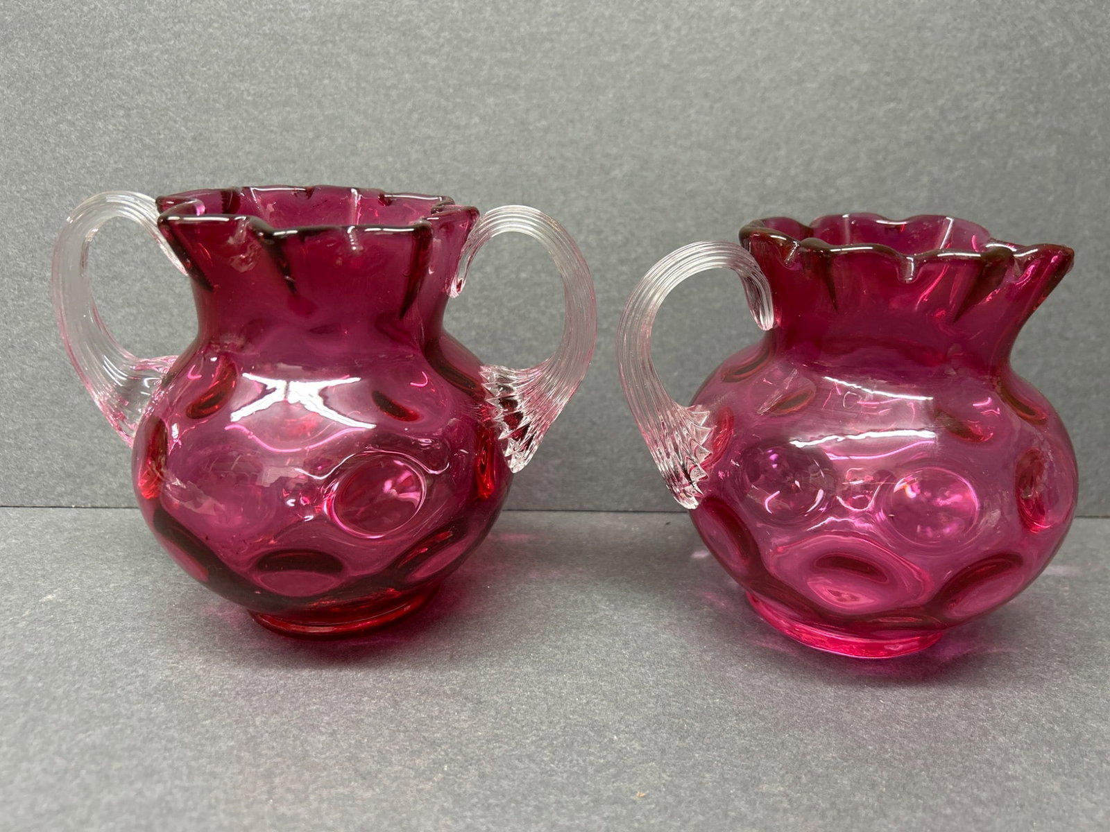 Cranberry Glass Creamers With Hobnail Design and Clear Handles: This lot includes a pair of cranberry-colored glass creamers with a hobnail texture and scalloped rims. Both feature clear glass handles and are approximately 3.5 inches tall, as indicated by the