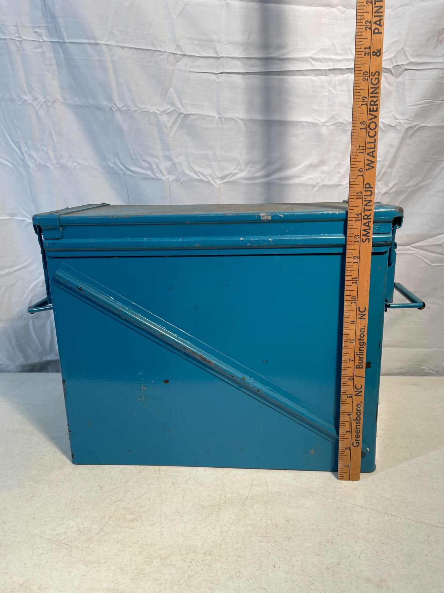Metal Storage Box with Hinged Lid and Interior Compartment: A rectangular metal storage box in blue featuring a hinged lid and carrying handles on both sides. The box measures approximately 12 inches in height and 18 inches in width, as indicated by the ruler