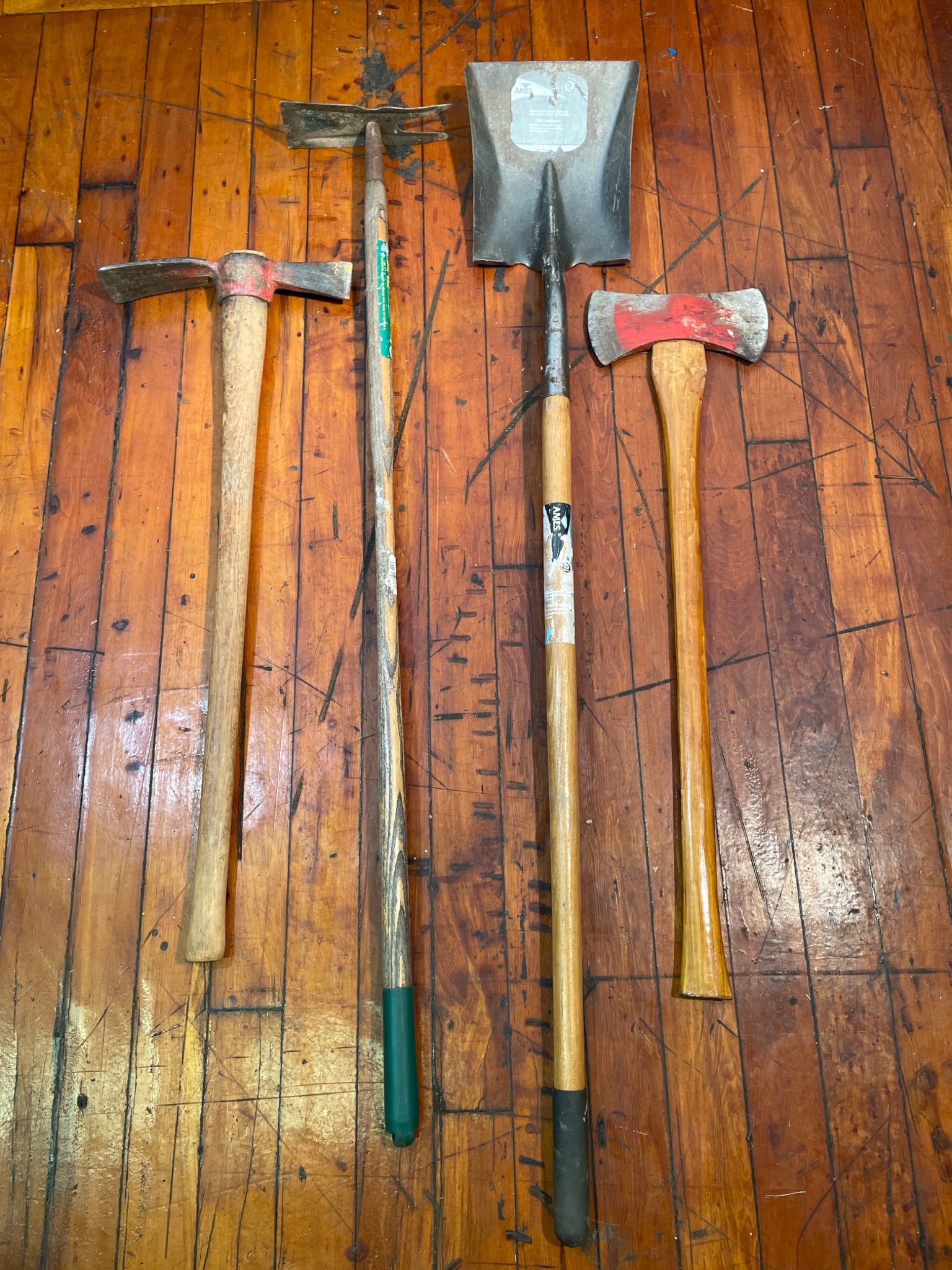 4 Assorted Yard Tools Including Axes, Hoe, and Shovel: This lot includes four yard tools: one shovel with a metal blade and wooden handle, one double-bit axe with a wooden handle, one pickaxe-hoe combination tool with a wooden handle, and one pickaxe