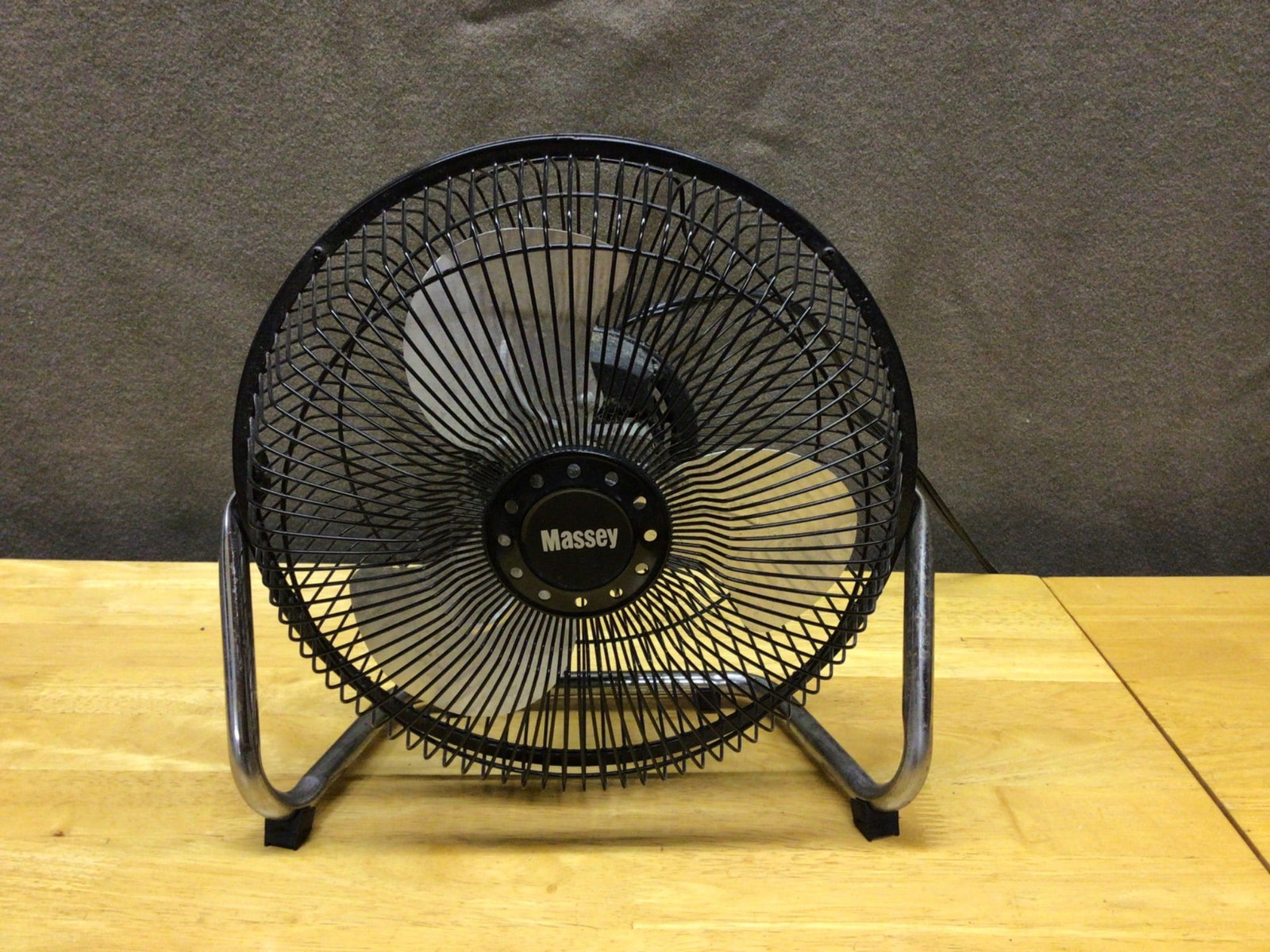 Massey 12-Inch Table Fan With Metal Grille and Adjustable Stand: Massey 12-inch table fan featuring a black metal grille and three-blade construction. The unit includes an adjustable metal stand and a rear-mounted rotary control for speed settings. Power cord