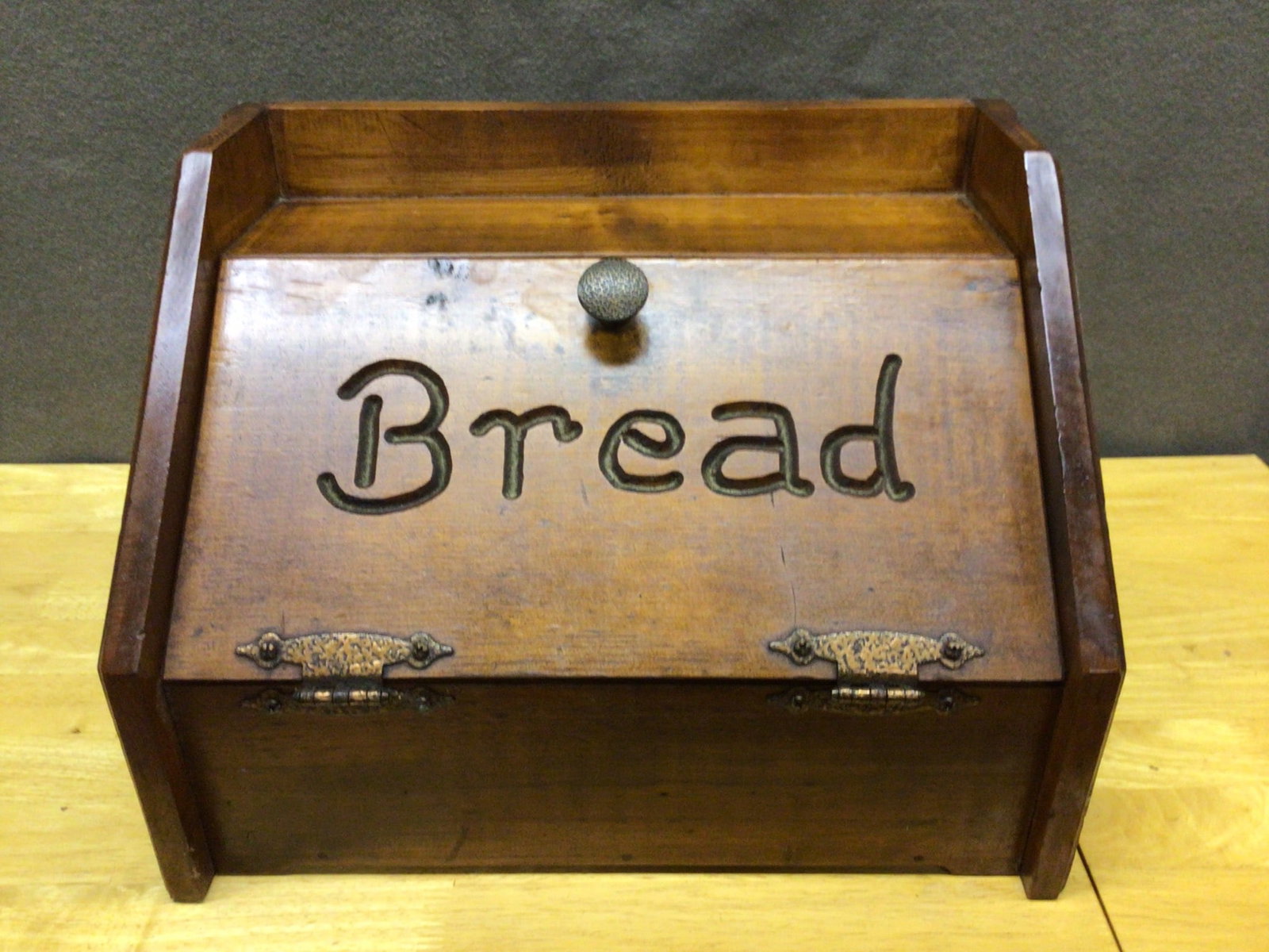 Wooden Bread Box With Hinged Lid and Carved 'Bread' Label: This is a wooden bread box featuring a slanted hinged lid with the word 'Bread' carved prominently on the front panel. The box is equipped with decorative metal hinges and a round knob for opening the