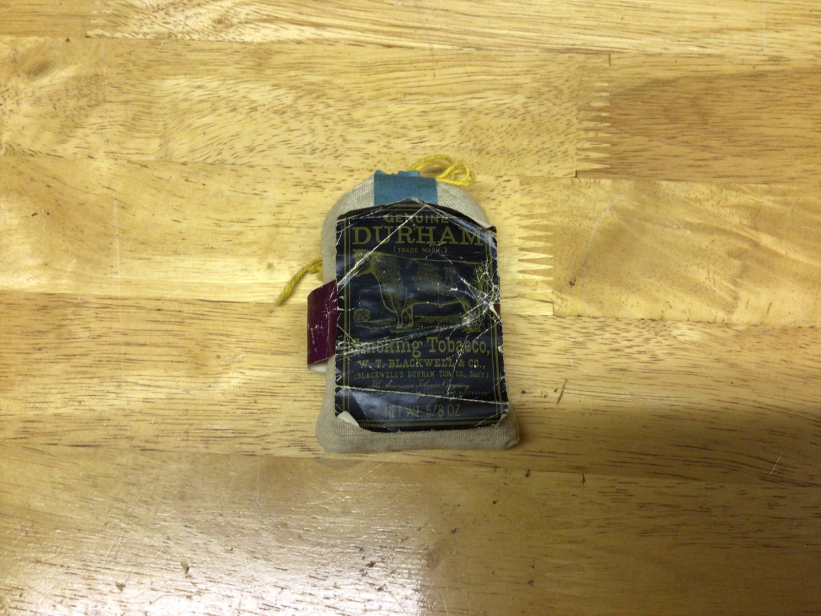 Durham Smoking Tobacco Packet with Label Featuring Bull Logo: A small fabric pouch containing Durham smoking tobacco. The original label with 'Bull Durham' branding is affixed but shows signs of wear. The pouch is measured at approximately 4.5 inches in height
