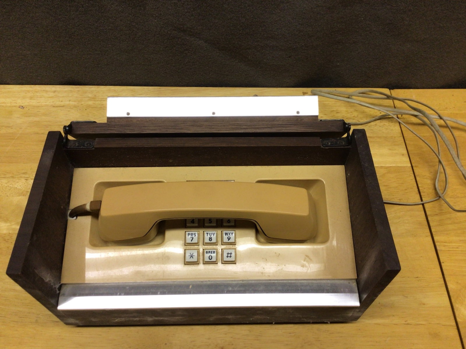 Wooden-Cased Telephone with Push-Button Dialing and Integrated Handset: This is a telephone encased in a rectangular wooden housing with a push-button keypad design. The case includes a drop-down lid for concealing the handset and keypad. The telephone features a beige