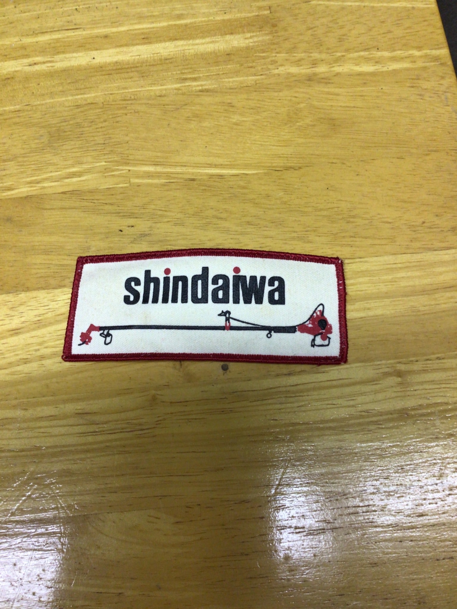 Shindaiwa Old Patch with Red Embroidered Border: This item is an old Shindaiwa patch featuring the company's name and a design of equipment. It measures approximately 4.5 inches in width and 2 inches in height. The patch has a red embroidered