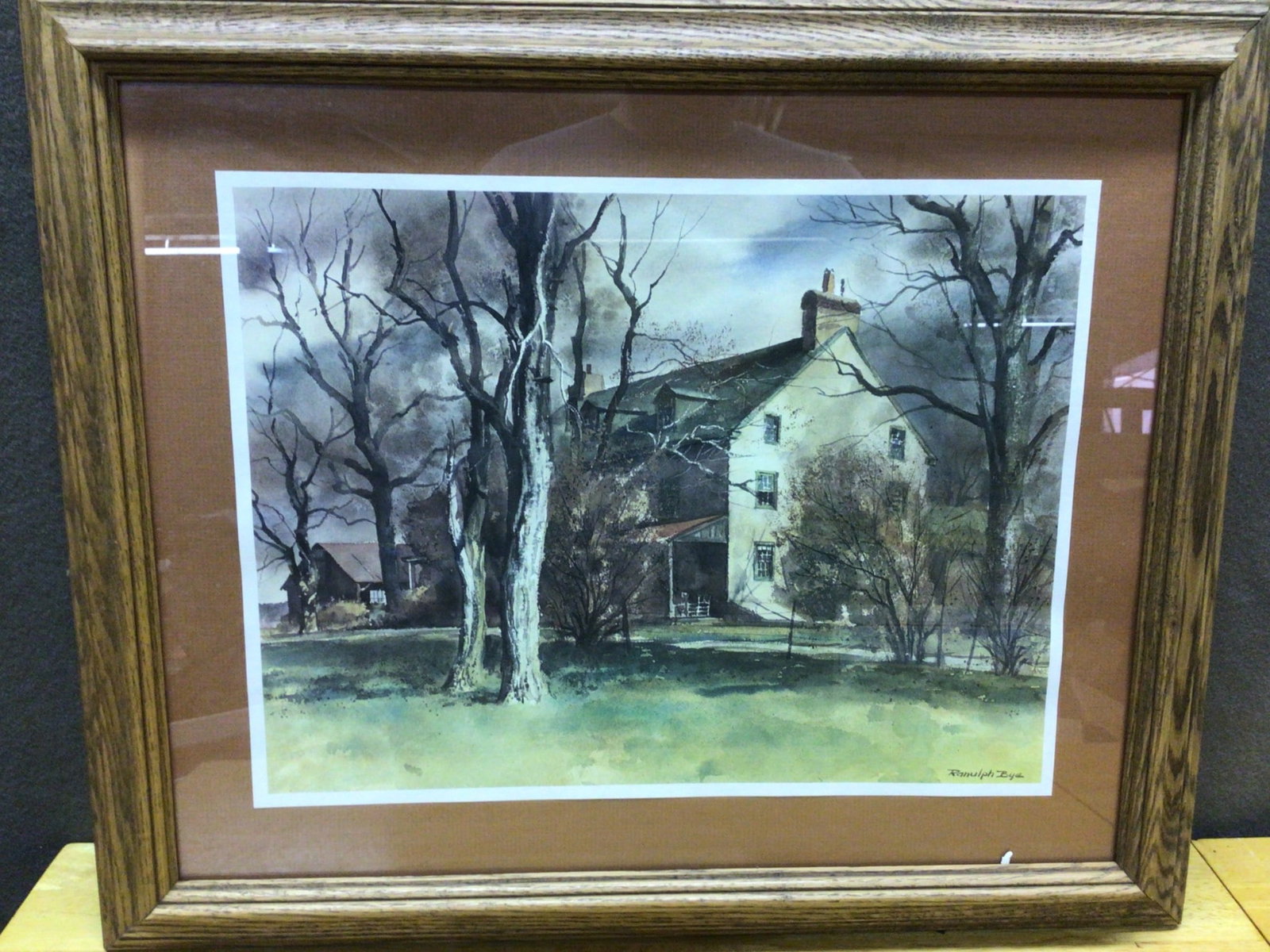 Signed Randolph Bye Framed Watercolor Print in Oak Frame: Framed watercolor print signed by Randolph Bye, featuring a serene home and trees landscape. The artwork is housed in an oak frame and measures approximately 20 inches by 16 inches (including the fram