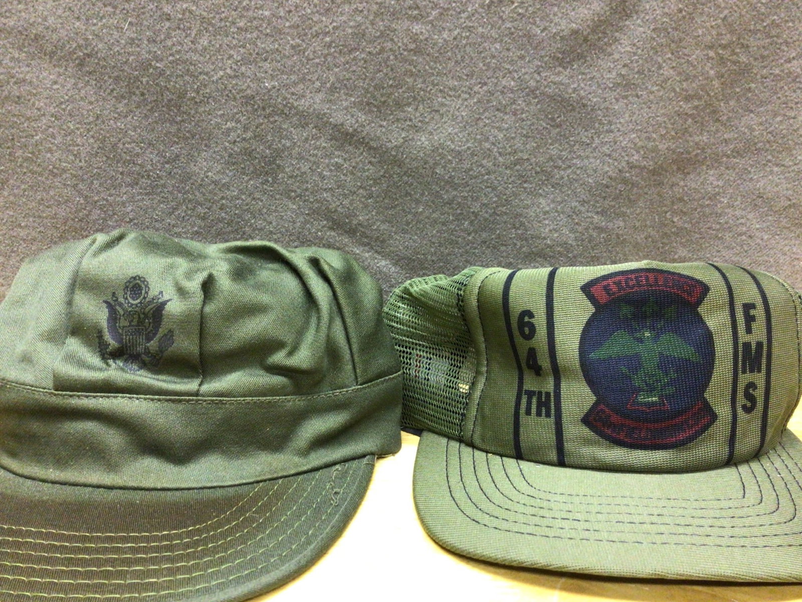 United States Military Headwear: Two Caps, Including 64th FMS Model: This lot includes two military-style caps. The first cap features an embroidered U.S. insignia on the front. The second cap showcases the 64th FMS (Field Maintenance Squadron) emblem on a mesh-back de