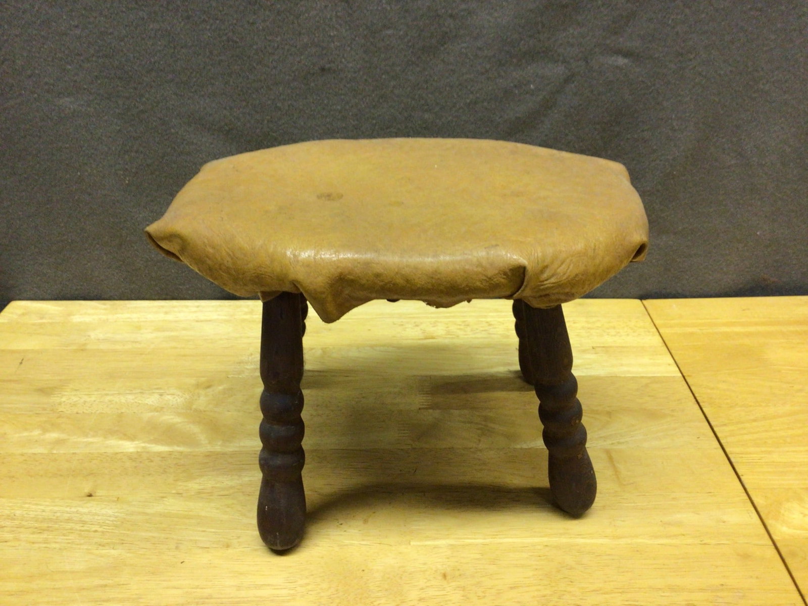 Handcrafted Footstool With Leather Upholstery and Wood Legs: This handcrafted footstool features a leather-covered top with rivet detailing and four wooden turned legs. The stool measures approximately 10 inches in height and 12 inches in diameter. The undersid