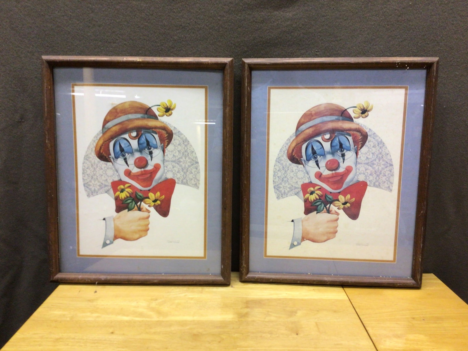 Framed Clown Prints By Tom Wood: This lot includes two framed prints by artist Tom Wood, each depicting a clown holding yellow flowers. The measurements, including frames, are approximately 22 inches tall by 18 inches wide. Frames