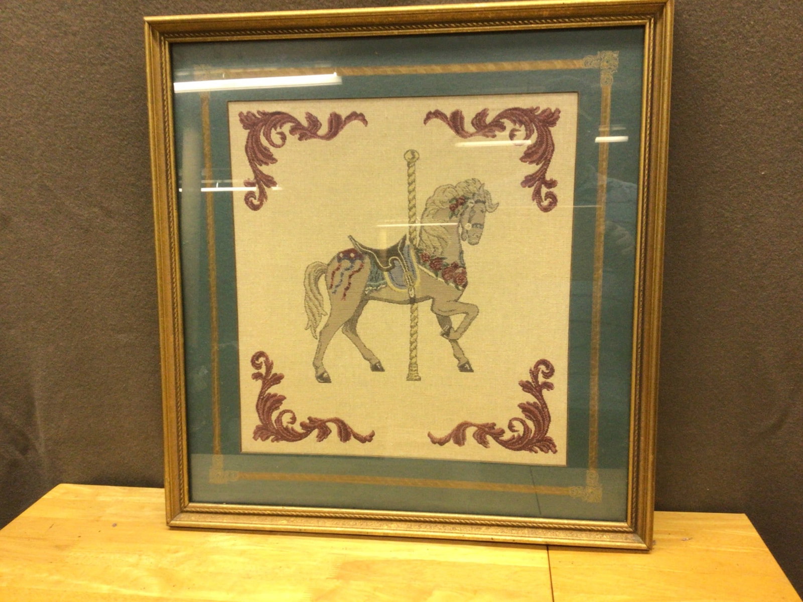 Handmade Embroidered Carousel Horse Artwork in Gilded Frame: Handmade embroidered artwork featuring a carousel horse motif, framed within a gilded wooden frame. The total frame dimensions are approximately 24 inches by 24 inches. The design incorporates ornate