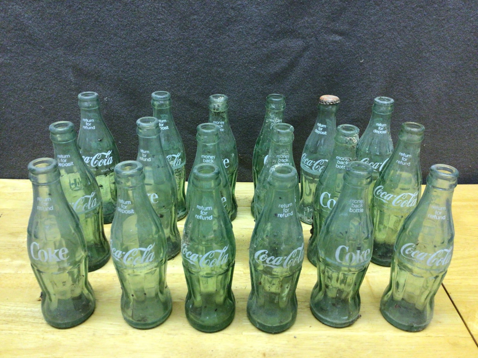 Empty Coca-Cola Glass Bottles With Embossed Text: This lot includes a collection of approximately 18 empty green Coca-Cola glass bottles. Most bottles feature embossed text such as 'Return for Refund' and 'Money Back Bottle.' One bottle includes a