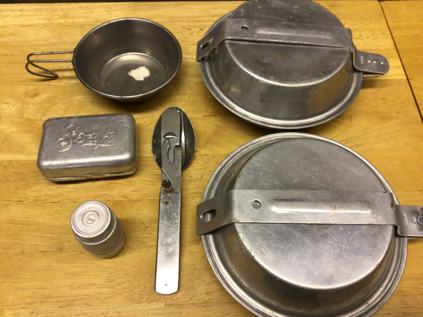 Aluminum Mess Kit with Utensils and Accessories, Never Used: This aluminum mess kit includes two pans with folding handles, a cup with a foldable handle, a combination spoon/fork utensil, a small container, and a tin case marked 'Soap'. The set appears