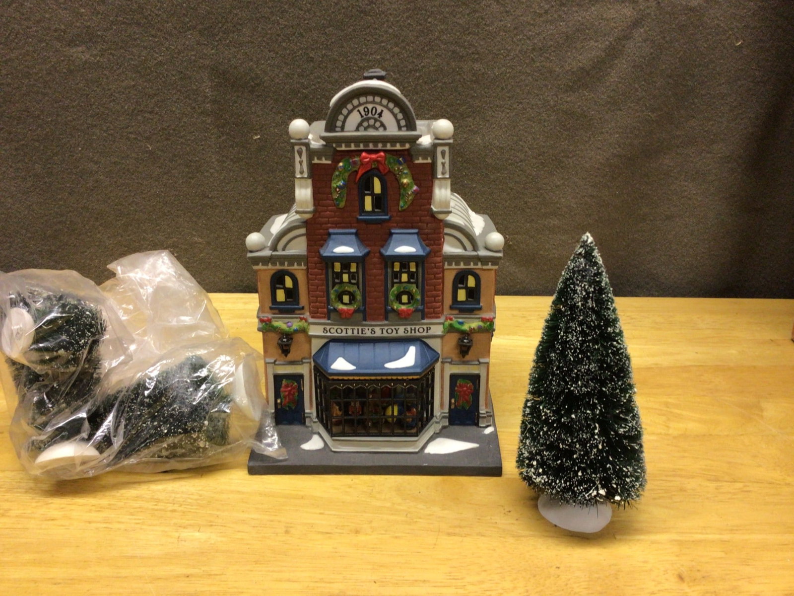 Scottie's Toy Shop Porcelain Lighted Building with Accessories: This is a Scottie's Toy Shop lighted porcelain building, part of a collectible series, as shown on its original packaging. The set includes the toy shop piece, additional bottle brush trees, and