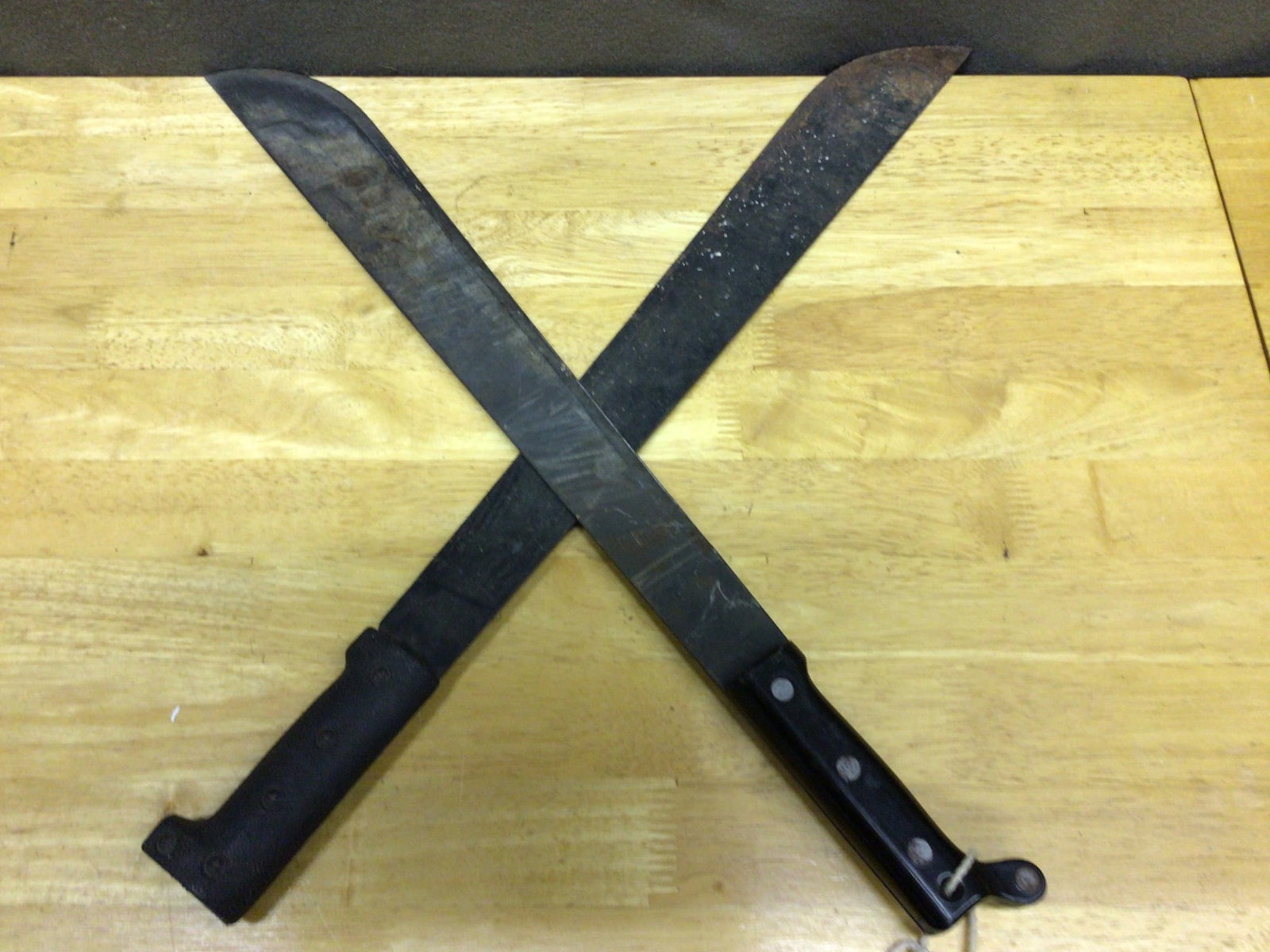 Two Machetes with Black Handles and Metal Blades: This lot consists of two machetes featuring black handles and solid metal blades. One machete has a molded black grip, while the other features a handle with visible rivets and an attached wrist