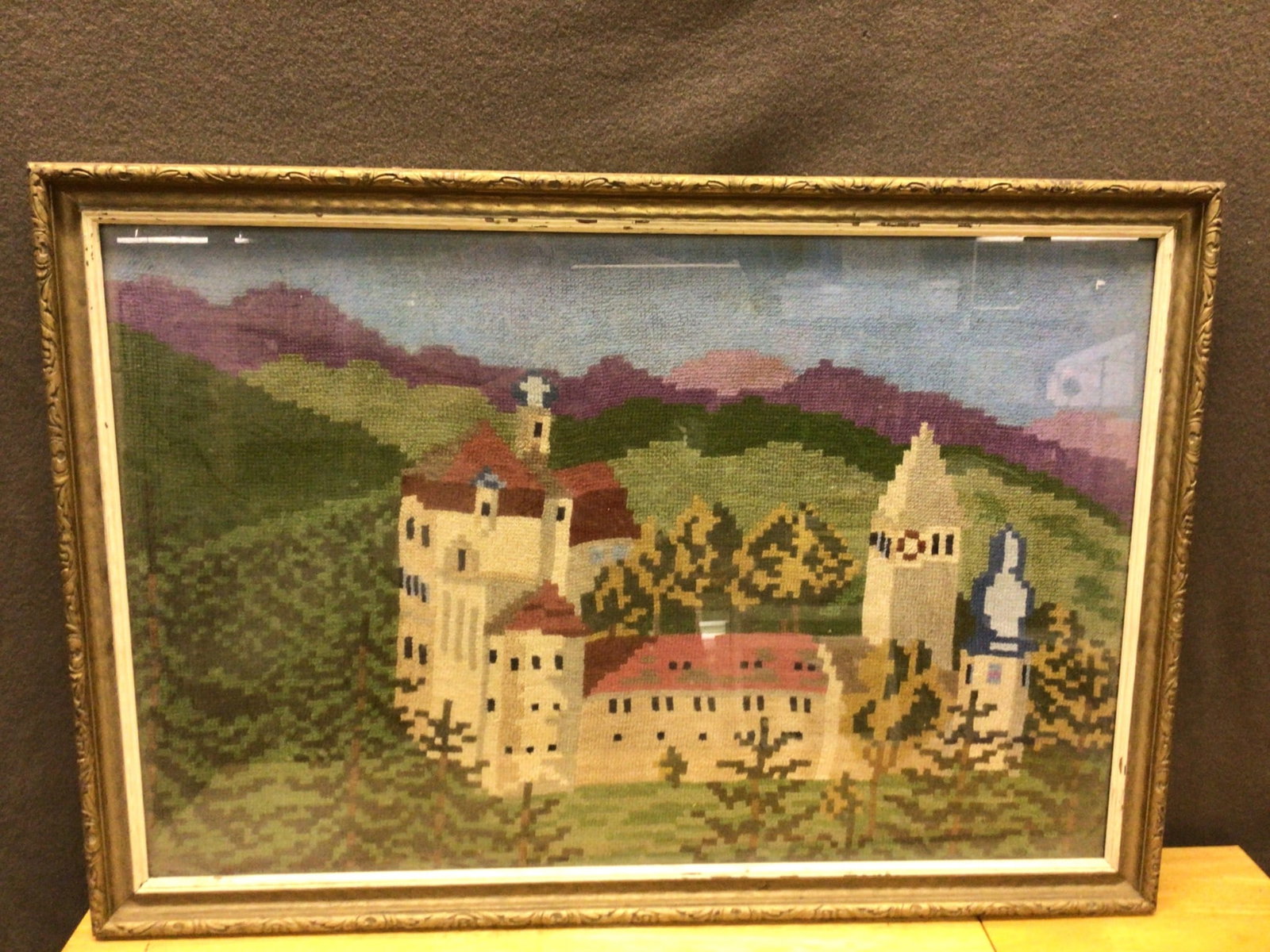 Framed Embroidered Landscape Depicting Castle Scene: A framed embroidered artwork depicting a castle scene with surrounding trees and mountains. The artwork is housed in a frame that measures approximately 28 inches by 20 inches. The embroidery