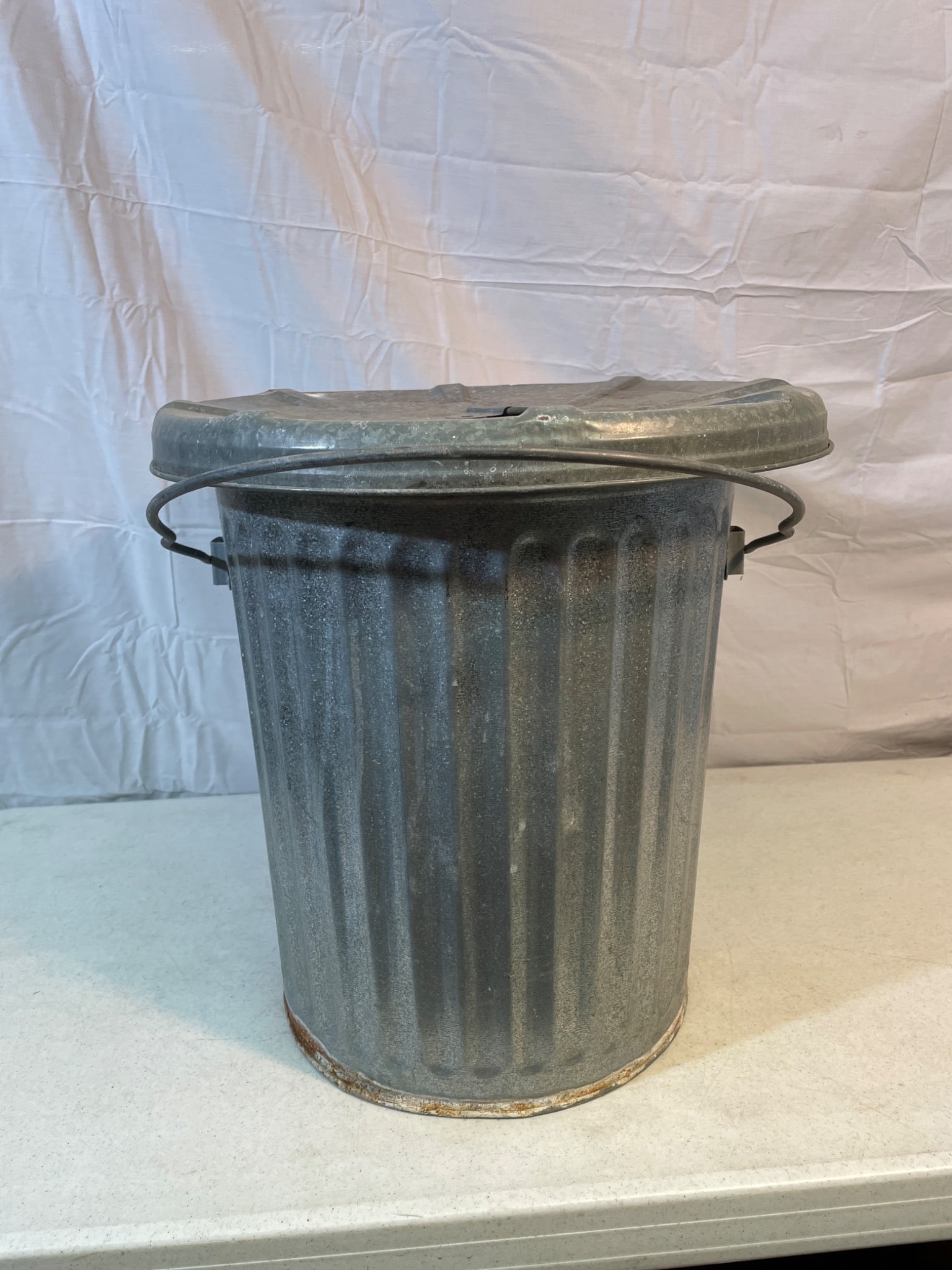 Metal Garbage Can with Lid and Handles, Possibly Galvanized Steel: This is a metal garbage can featuring a removable lid with a central handle and two side carrying handles. The construction suggests galvanized steel. It shows signs of wear, including rust and