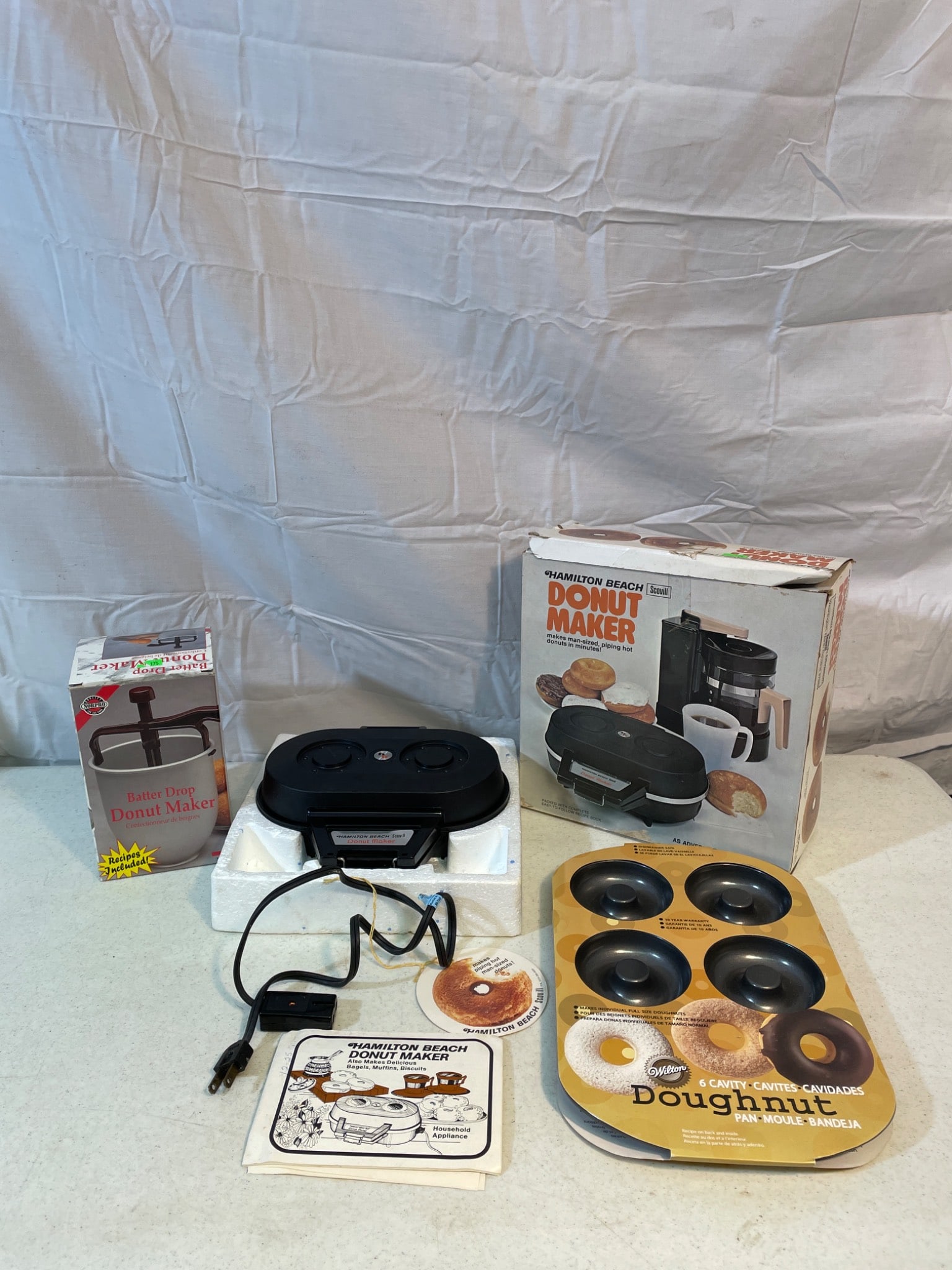 Donut Maker Set Including Donut Maker, Wilton Doughnut Pan, and Accessories: This lot includes a donut maker appliance (tested and it works), its original box, a 6-cavity Wilton Doughnut Pan, and additional accessories as shown. The donut maker appears complete with its power