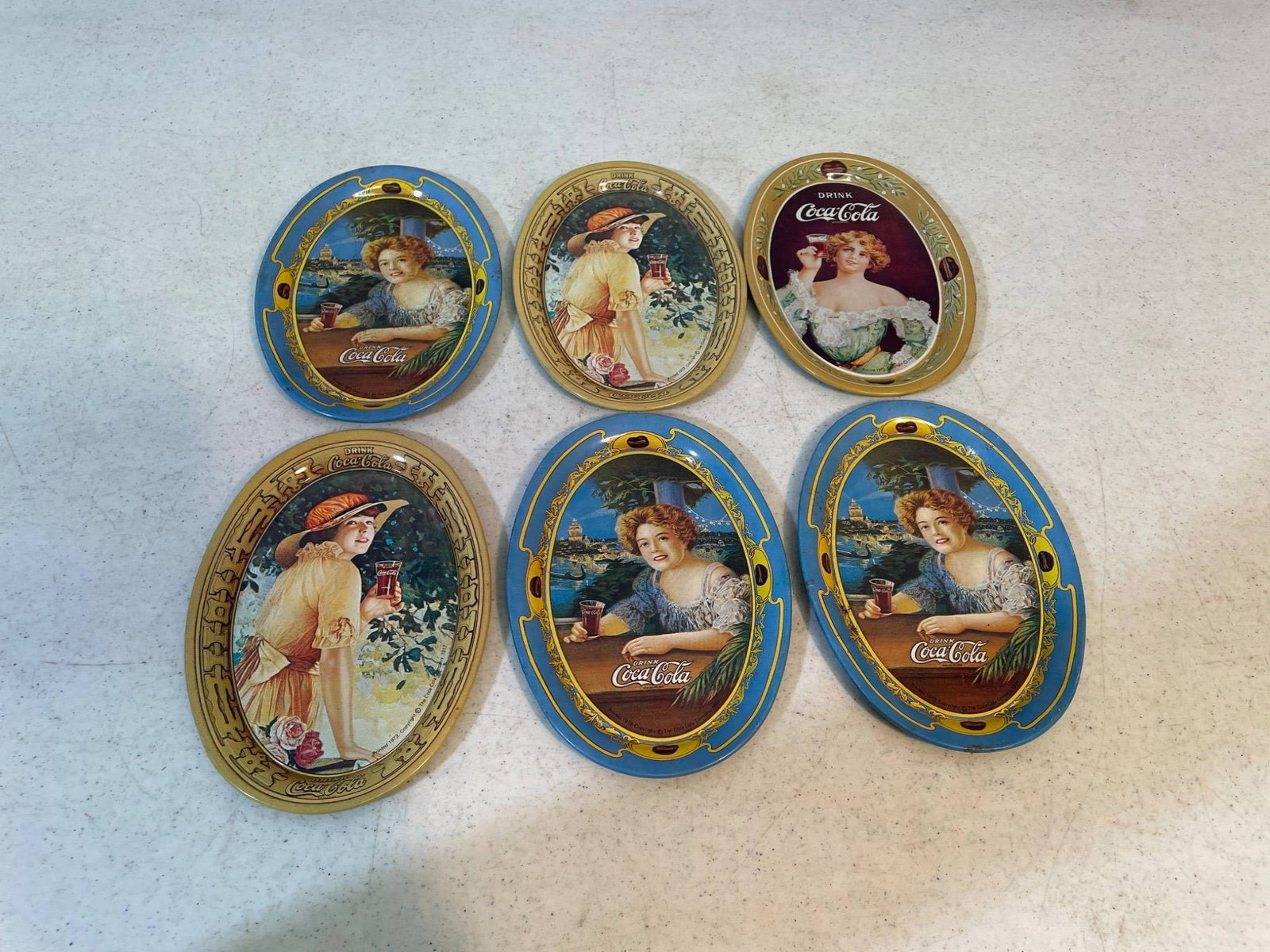 6 Coca-Cola Decorative Oval Metal Trays with Vintage Artwork: This lot includes six oval-shaped Coca-Cola decorative metal trays featuring vintage-style artwork of women. The trays display vibrant illustrations alongside Coca-Cola branding. The backs are plain
