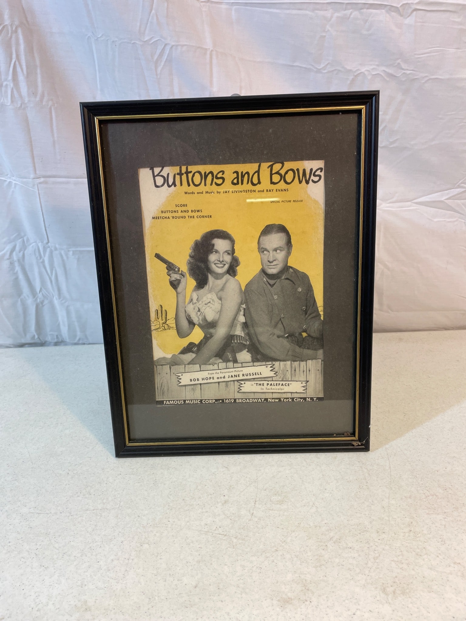 Framed 'Buttons and Bows' Sheet Music with Black Frame: This is a framed piece featuring the sheet music cover for 'Buttons and Bows.' The ad features artwork of two individuals in 1940s-style attire. The black frame includes a hanging mechanism on the bac