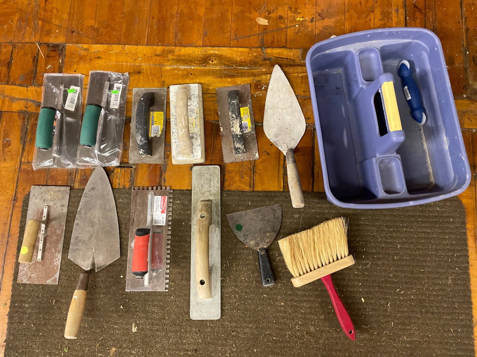 Various Trowels, Brushes, and Plastic Storage Bin: This lot includes a variety of construction tools such as trowels (smooth, notched, and pointed), rubber grip handles, a block brush, and a plastic storage bin. Some items appear to have retail sticke