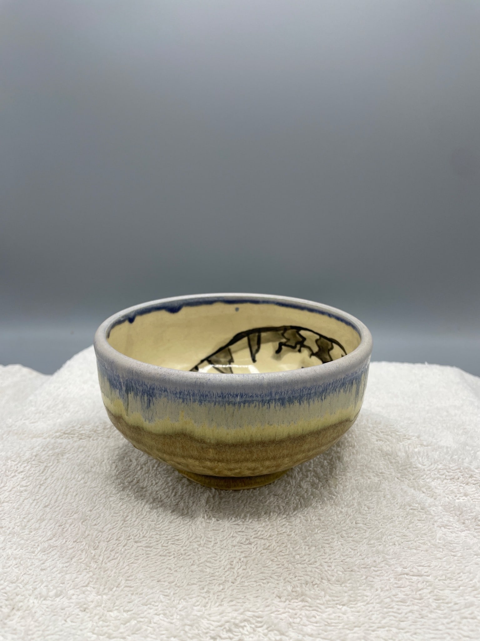 Handcrafted Ceramic Bowl With Painted Cat Design and Stamped Mark: A handcrafted ceramic bowl featuring a whimsical cat design with a party hat painted on the interior. The exterior displays a mottled beige and blue glaze transitioning to unglazed ceramic at the