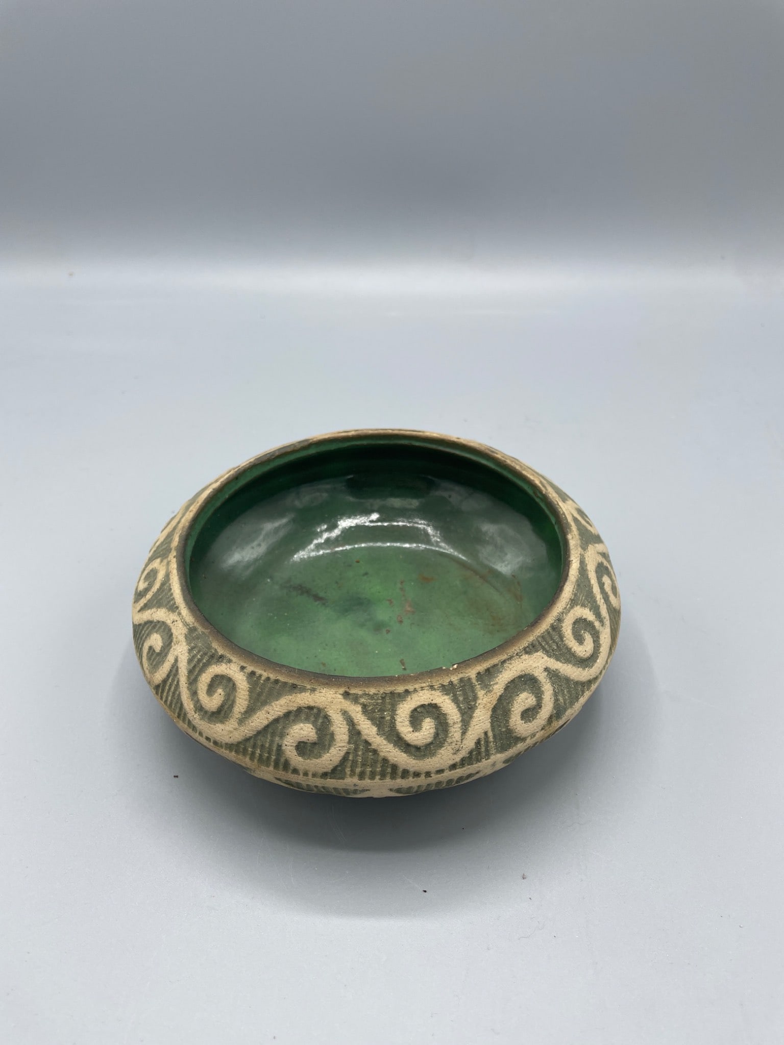 Stoneware Bowl with Green Glaze and Carved Swirl Motif: A decorative stoneware bowl featuring a green glazed interior and carved swirl motifs around the outer edge. The underside shows geometric carvings. Dimensions are approximately 2.75 inches in height