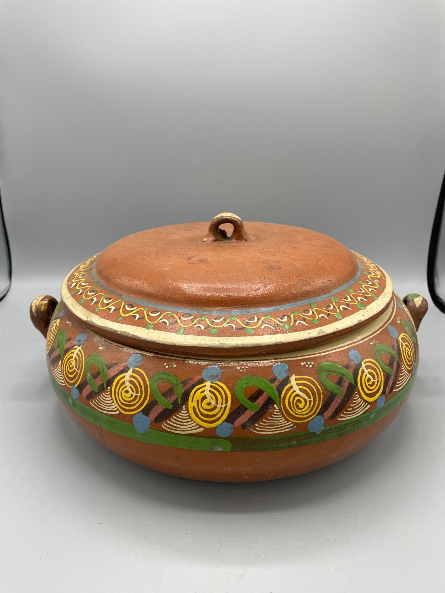 Hand-Painted Earthenware Pot with Lid and Handles: A round earthenware pot featuring hand-painted geometric and abstract patterns in various colors. The pot includes a fitted lid with a loop handle and two side handles for portability. Approximate