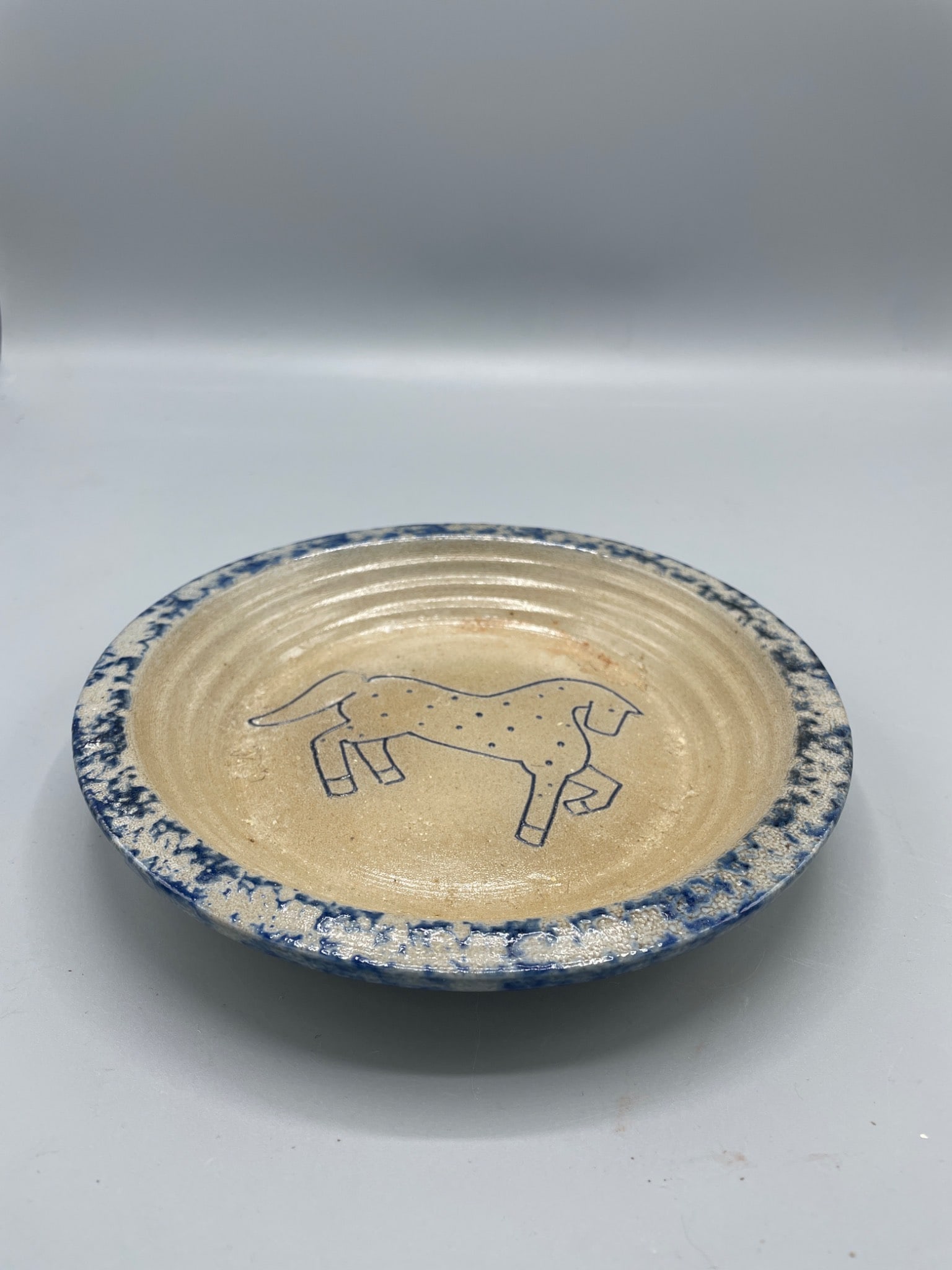 1992 Signed Ceramic Plate with Blue and Beige Finish Featuring Horse Motif: A hand-thrown ceramic plate featuring a blue and beige color palette with a horse motif on the face. The bottom is signed 'E. Mead 1992' alongside a star symbol. The plate measures approximately 6