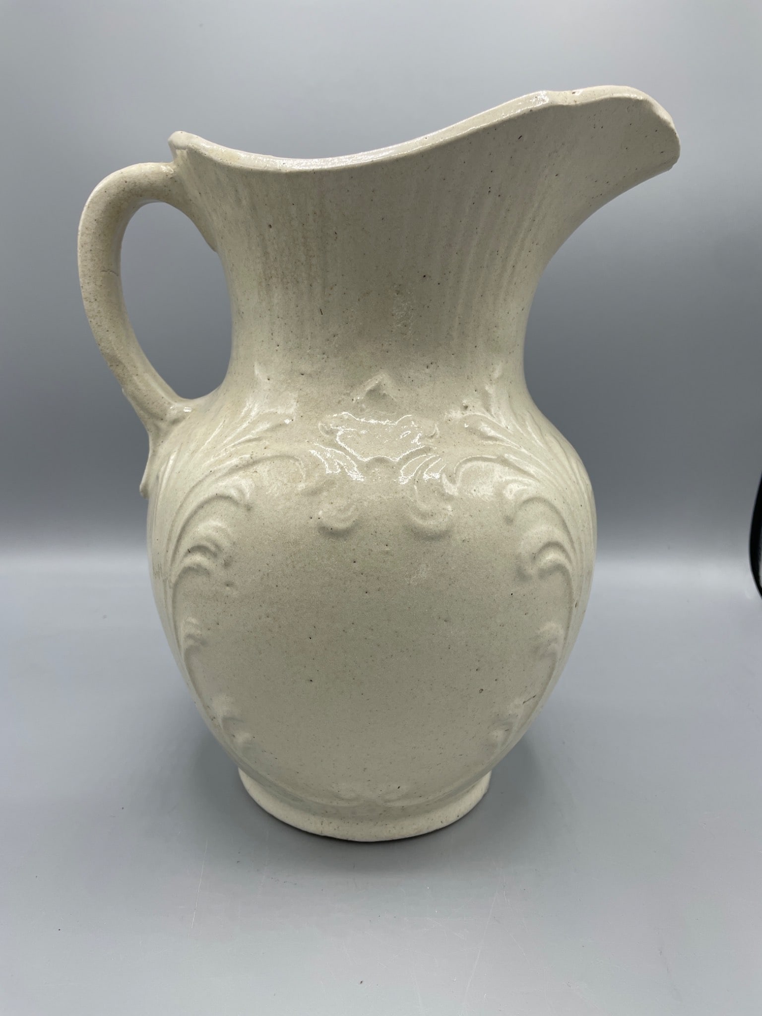 White Ceramic Pitcher With Relief Decoration and Flared Spout: This is a white ceramic pitcher featuring a textured relief design and a curved handle with a flared spout. The height measures approximately 12 inches. The base shows typical wear. The item exhibits