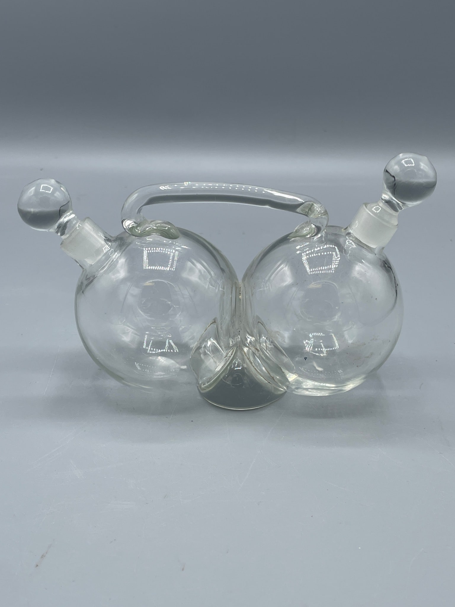 Dual-Chamber Glass Cruet With Stoppered Lids and Swing Handle: Glass dual-chamber cruet featuring two spherical containers with individual stoppers and a swing-style glass carrying handle. Each chamber has a ground opening for a secure seal with the stoppers. Ove