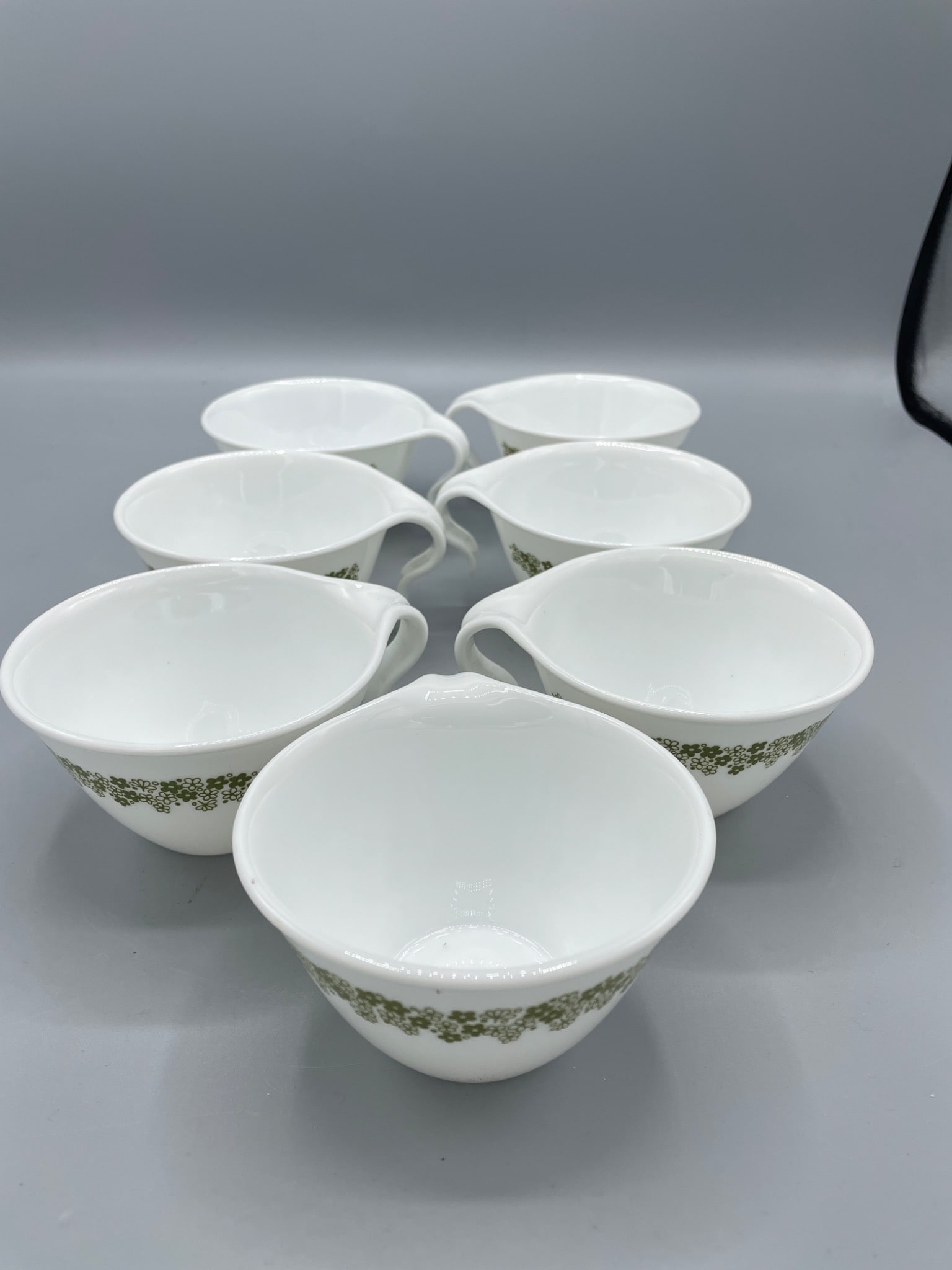 7 Corelle by Corning Mugs with Green Pattern: This is a set of 7 Corelle by Corning mugs, featuring a green floral or vine-like pattern along the rim. The bottom of each mug is marked 'Corelle by Corning, Corning, NY, USA - Not For Broiler Or
