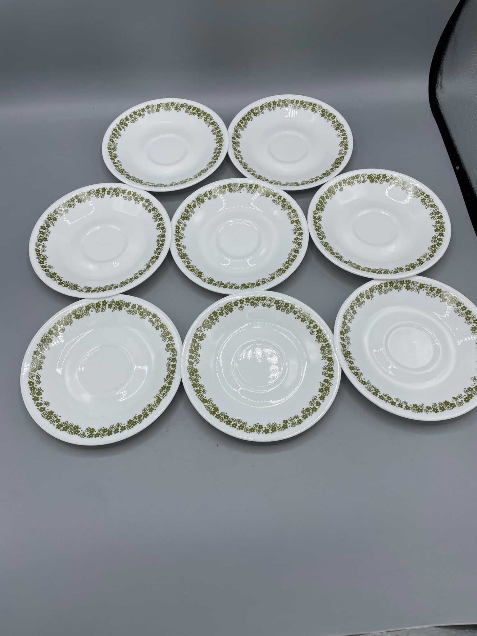 17 Corelle by Corning Saucers, Green Flower Border Pattern: This lot includes 17 matching saucers by Corelle, manufactured by Corning in Corning, N.Y., USA. Each saucer features a green flower border pattern. The underside is marked with the manufacturer's