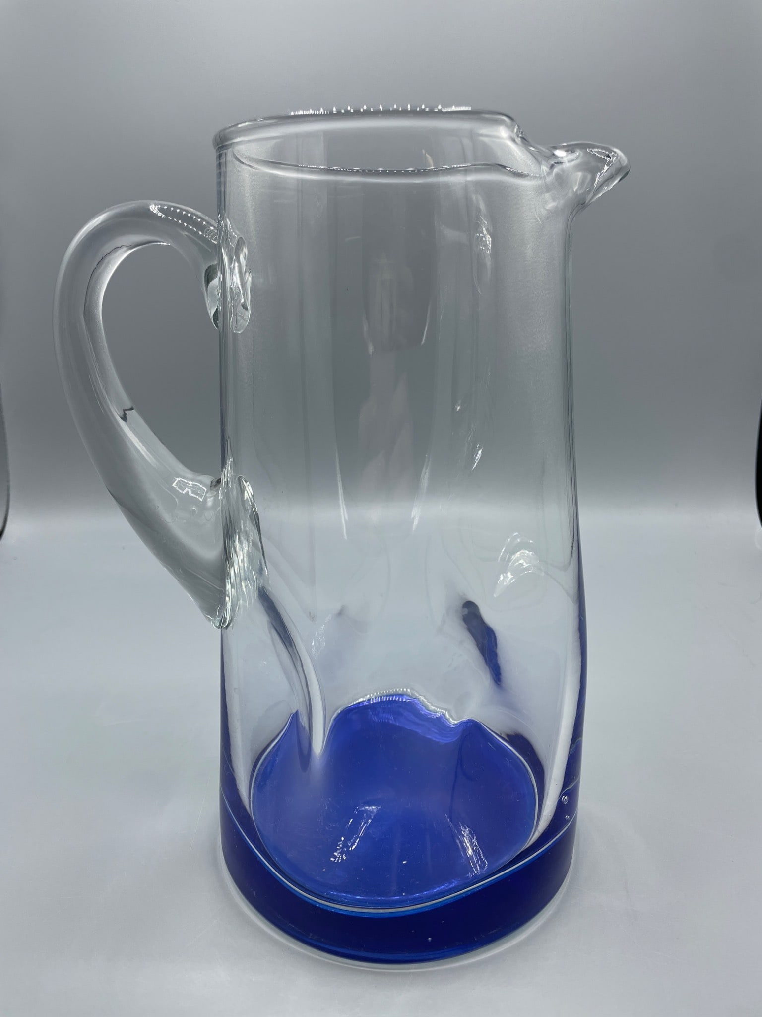 Clear Glass Pitcher With Blue Base And Handle: This is a clear glass pitcher featuring a blue-colored base and an integrated handle. It has a spout for pouring liquids and appears to measure approximately 10 inches in height. The item's condition