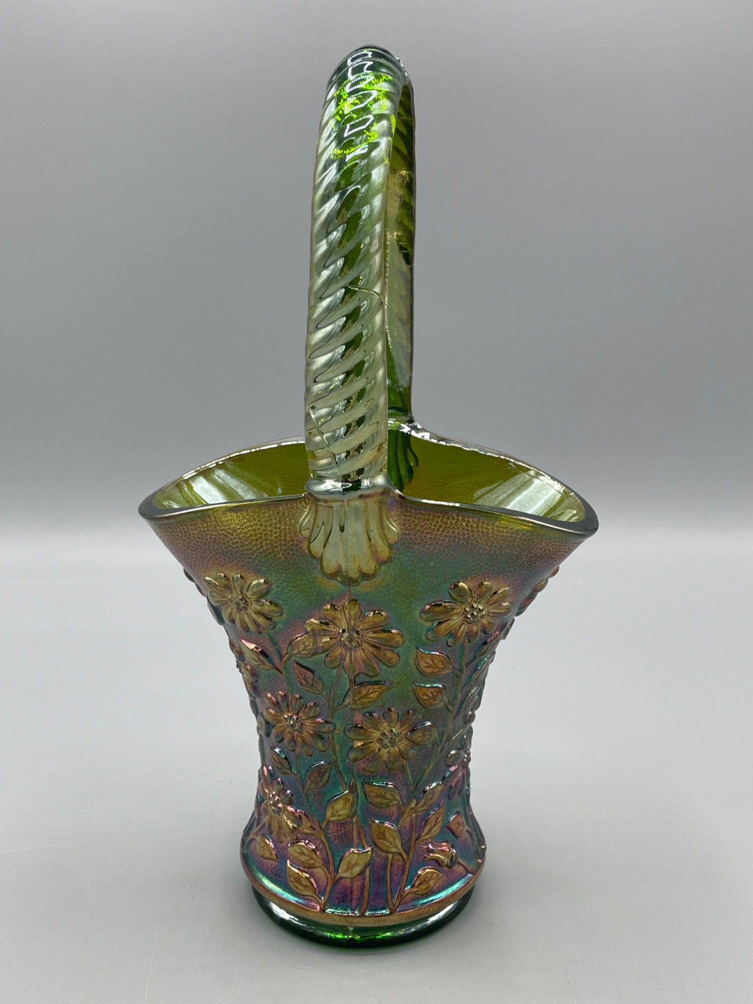 Green Iridescent Glass Basket With Twisted Handle and Floral Design: A green iridescent glass basket featuring a twisted handle design with floral embossing around the body. The basket exhibits a textured exterior, intricate details, and a vibrant iridescent finish.