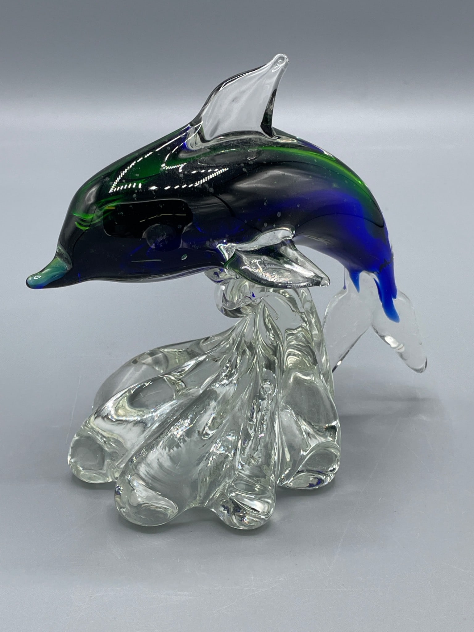 Glass Dolphin Figurine on Textured Base with Blue and Green Accents: Glass dolphin figurine featuring blue and green accents transitioning to clear glass, mounted on a textured base designed to resemble water splashes. The piece is approximately 6 inches tall