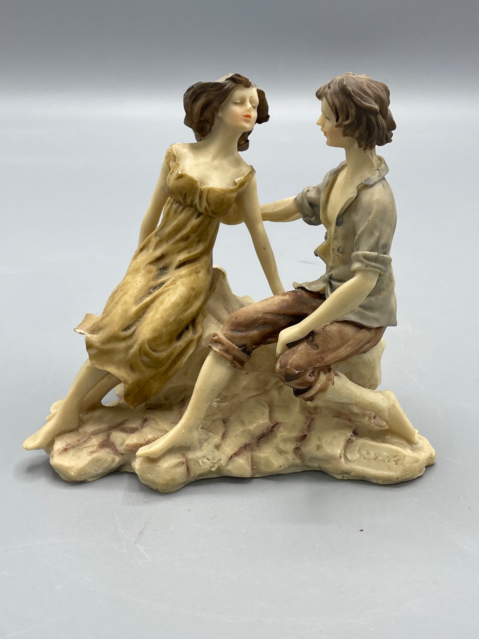 Hand-Painted Figurine of Man and Woman Sitting on Rock: Figurine depicting a seated man and woman on a textured rock base. Piece is hand-painted with naturalistic tones. Material appears to be a type of molded resin. No discernible maker's marks or model